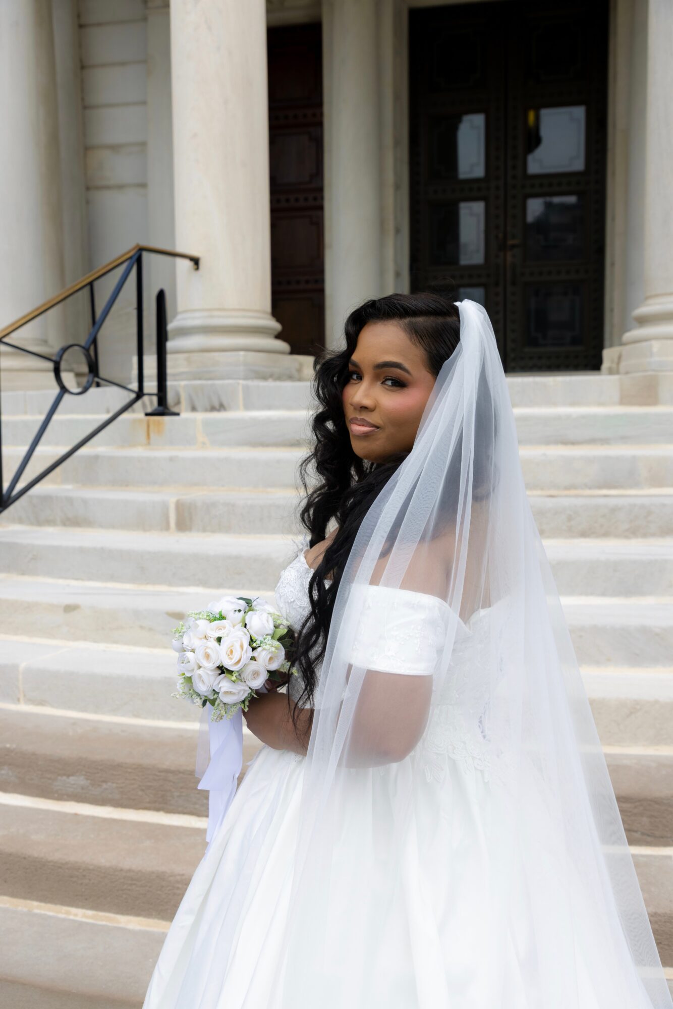 Bride in wedding dress holding bouquet standing on steps outside building with columns and large door