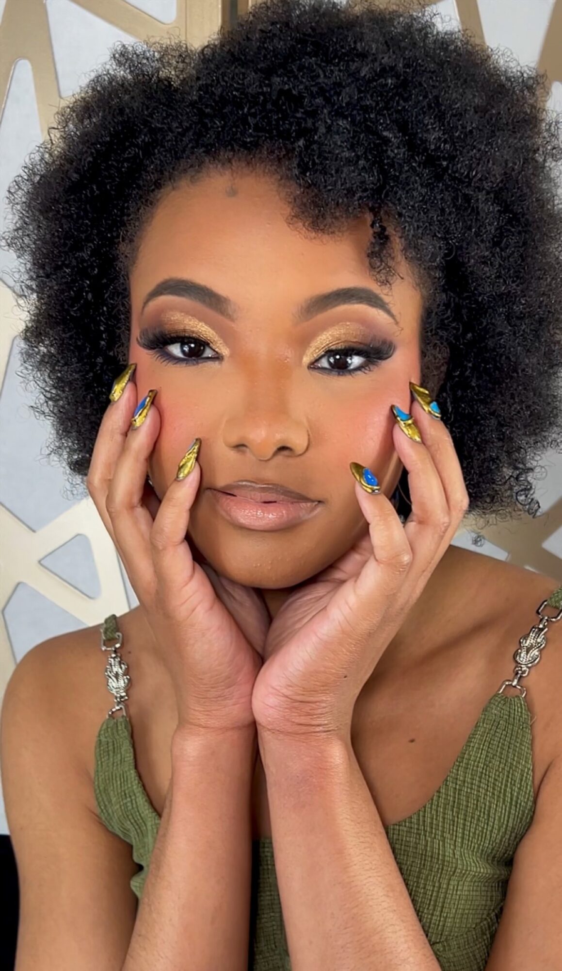 Close-up of a woman with natural curly hair, makeup, and colorful nails, touching her face with both hands.