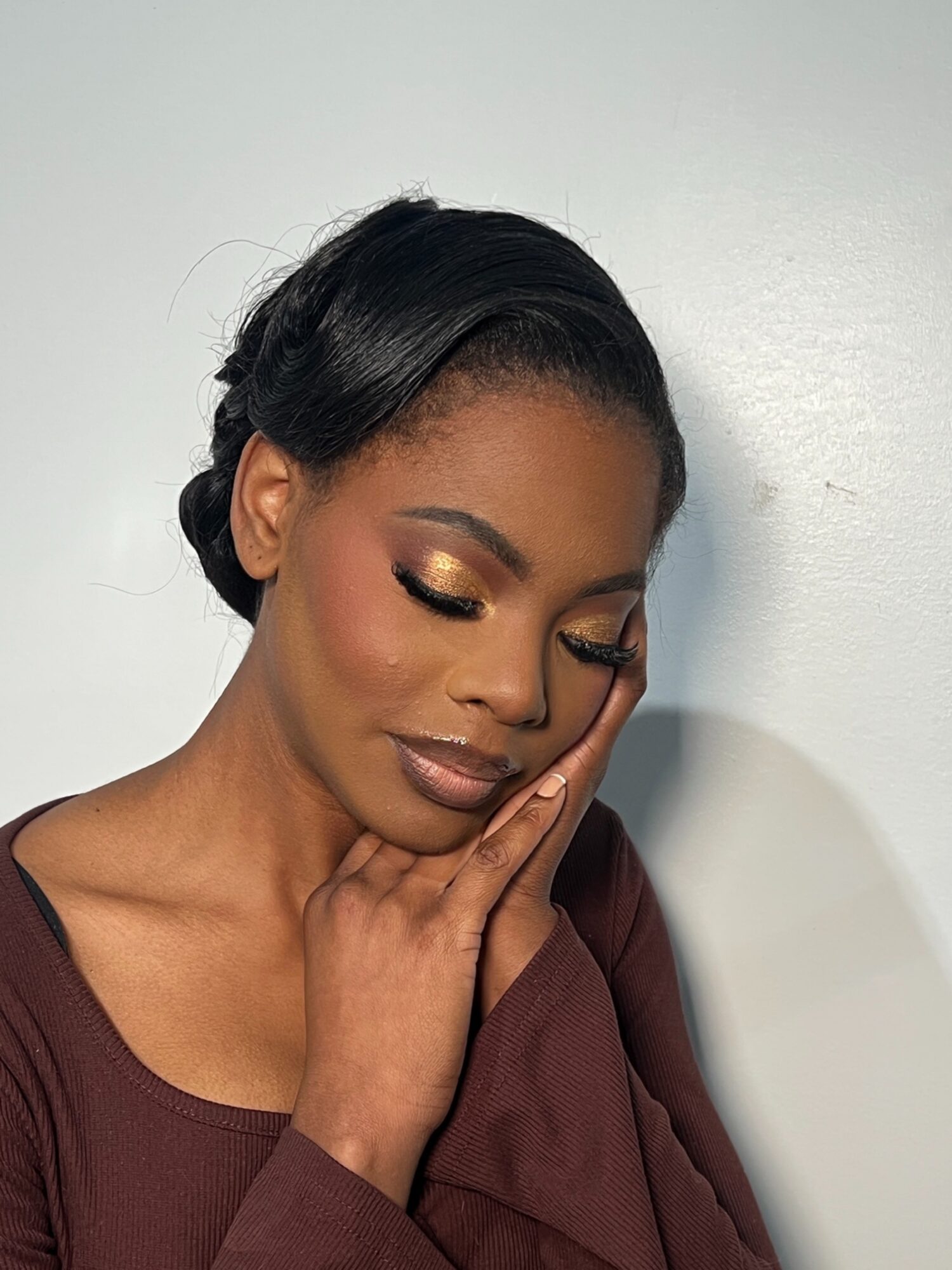 Woman with makeup and styled hair gently touching her face, eyes closed, against a plain background.