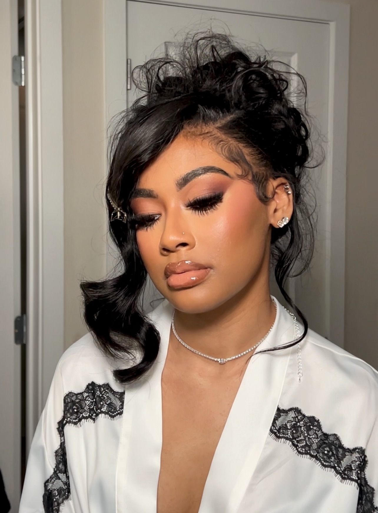 Young woman with curly hair styled in an updo, makeup, and a white top with black lace details, standing indoors.