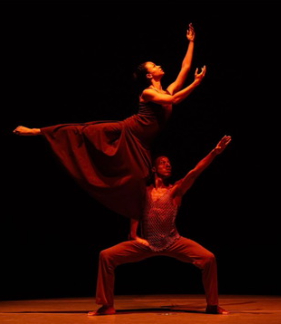 Two dancers perform on stage, one balancing on the other's shoulders, with a dark background.