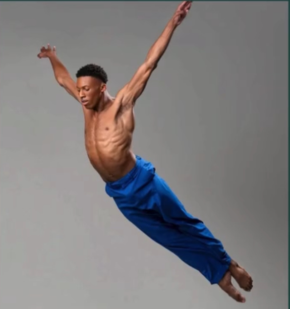 Young man with a muscular build jumps with arms and legs extended, wearing blue pants, against a plain background.