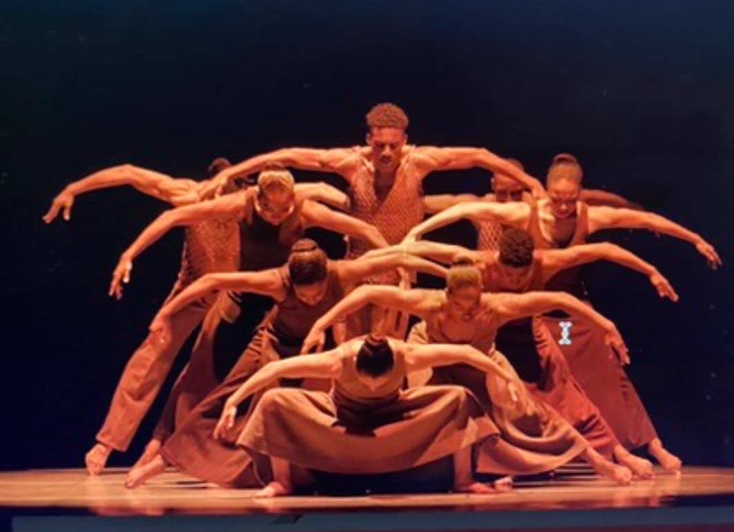 Group of nine dancers in a formation, stretching arms outward, performing on stage with dark background.