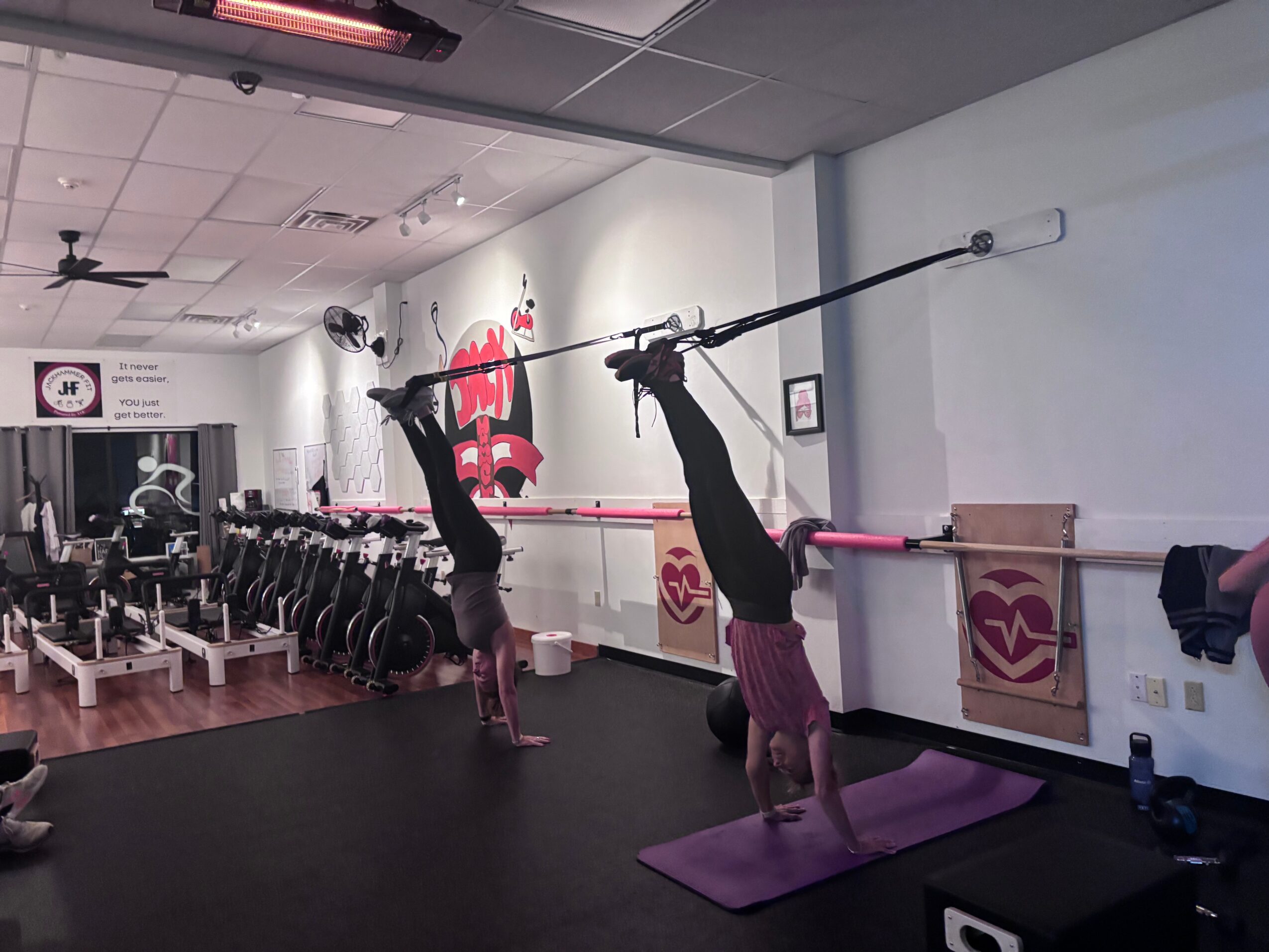 Two people perform handstands against a wall in a fitness studio, with exercise equipment and a mural in the background.