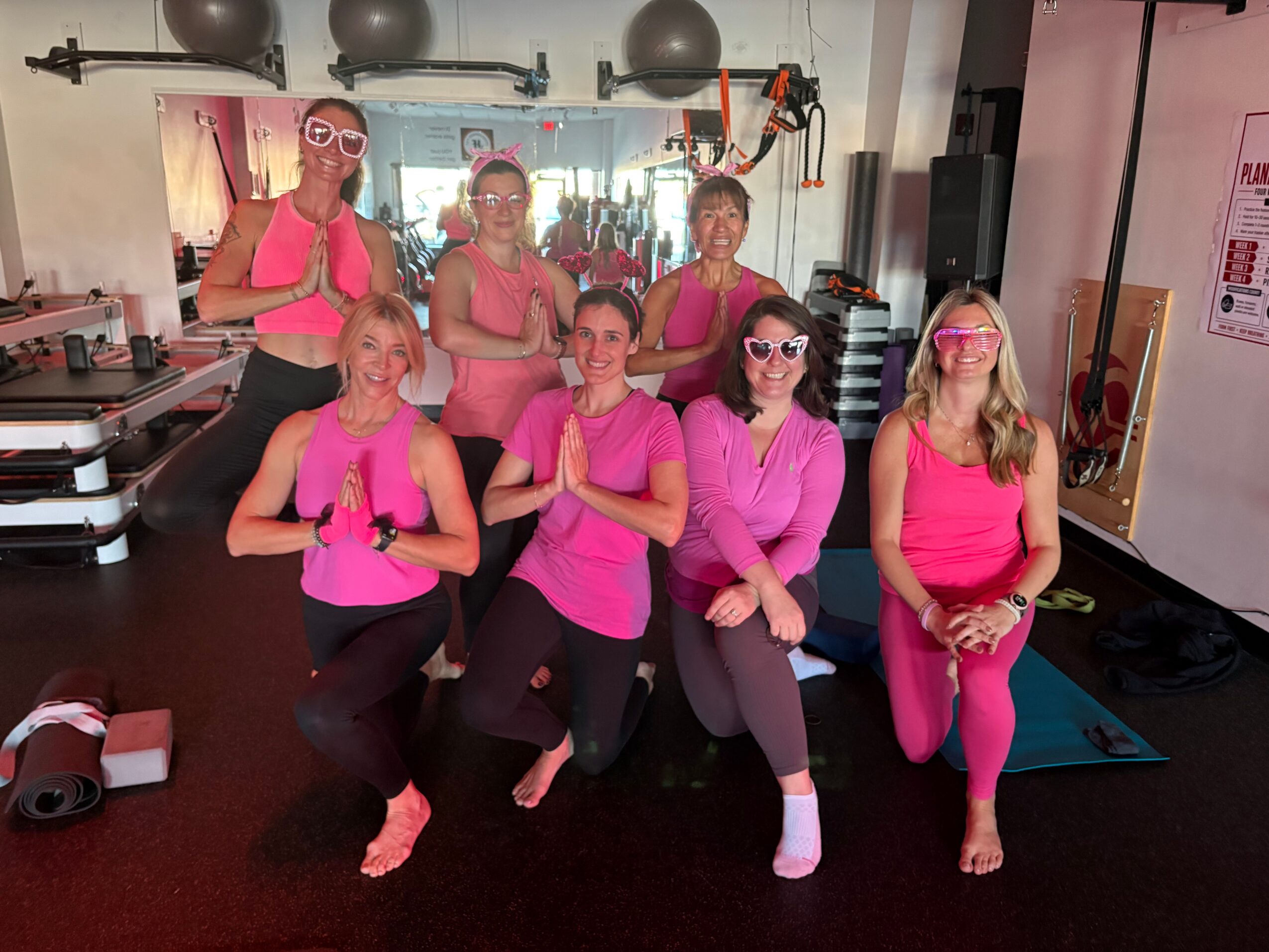 Group of seven women in pink workout clothes posing in a gym, some with hands in prayer position, smiling.