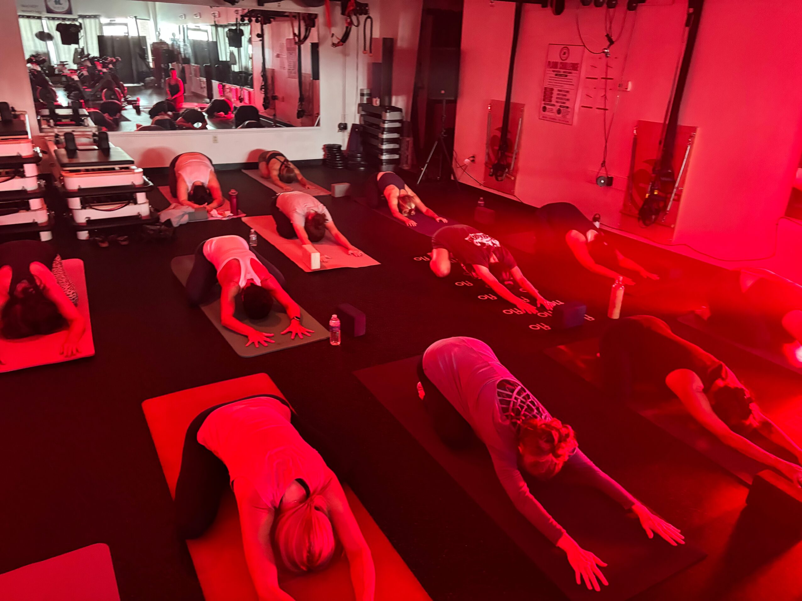 People practicing yoga in a fitness studio with mirrors, mats, and weights, illuminated by red lighting.