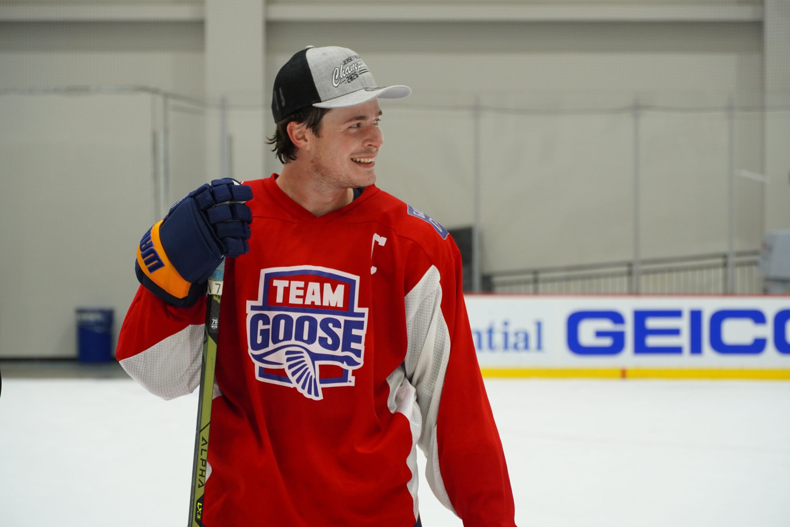 Young man in hockey gear smiling, wearing a red jersey with 'TEAM GOOSE' logo, on ice rink.