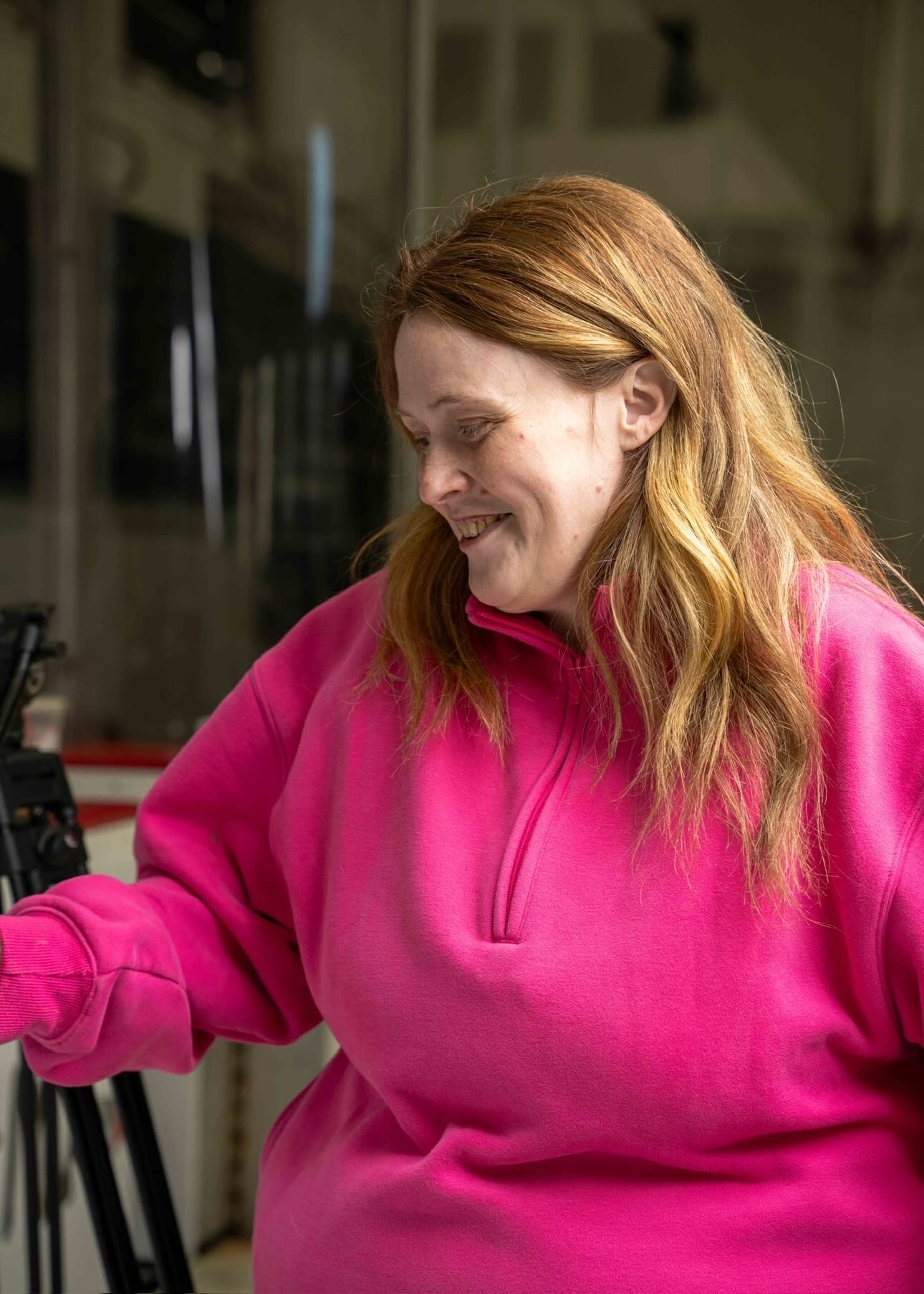 Woman with long red hair smiling, wearing a pink fleece, in an indoor setting.