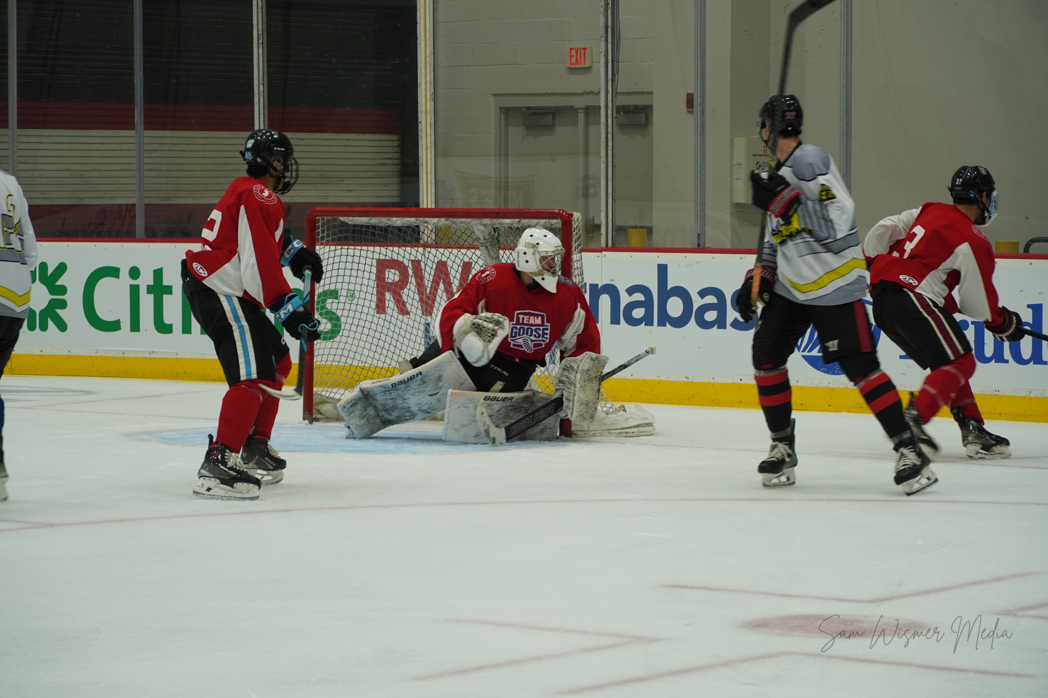 Hockey players in action near the goal, with a goalie in red and white gear making a save, and other players nearby.