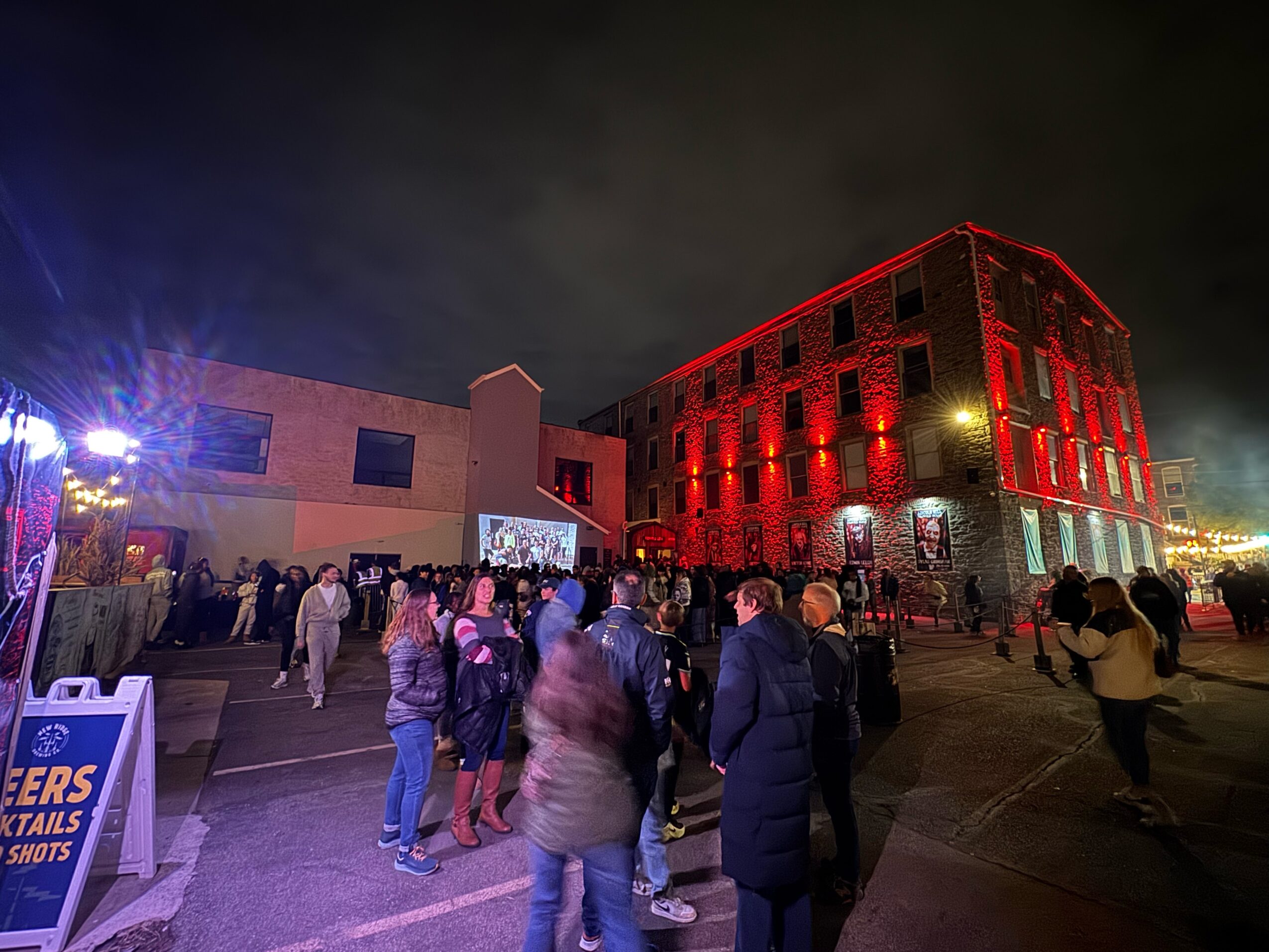 Night scene with a crowd of people outside a building illuminated in red lights, with a dark sky and some streetlights.