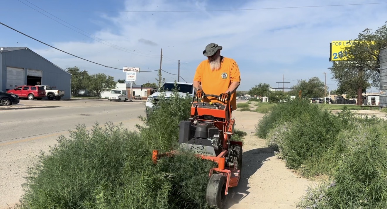 Person operating a lawn mower on a sidewalk with bushes, wearing an orange shirt and hat, in a commercial area.