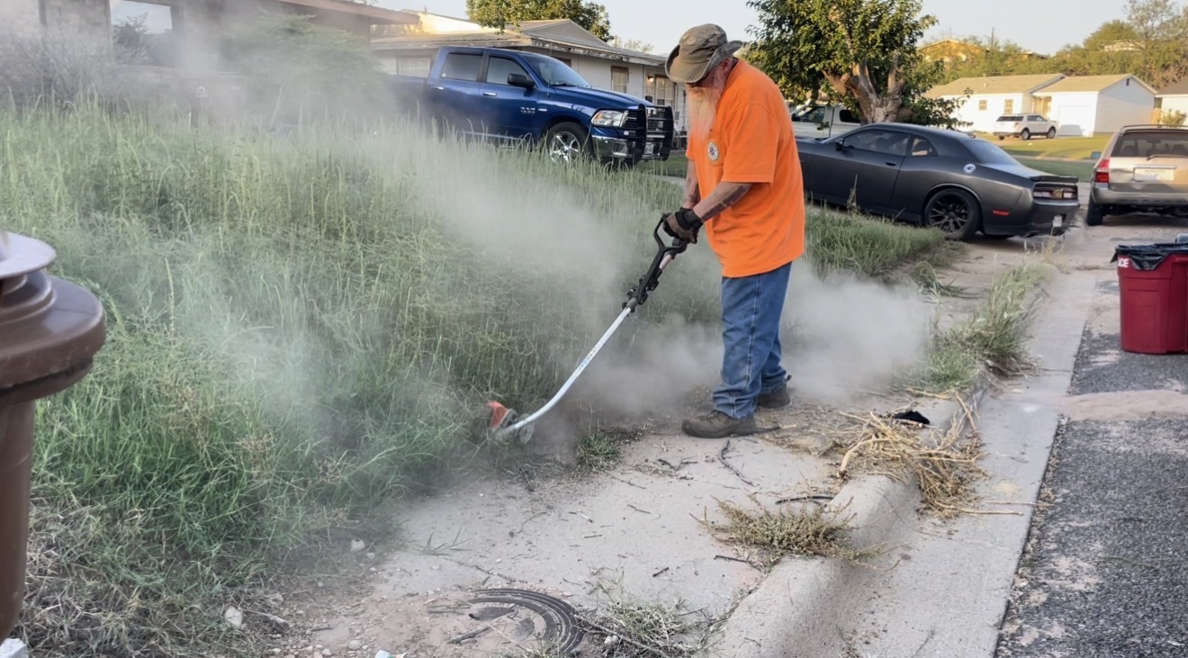 Person in orange shirt using a weed trimmer on overgrown grass by sidewalk, with smoke or dust nearby.