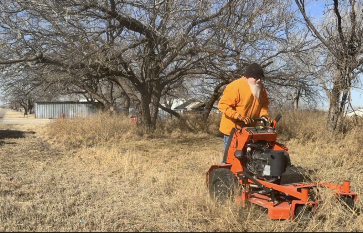Person in orange jacket operating a lawn mower in a field with trees and a building in the background.