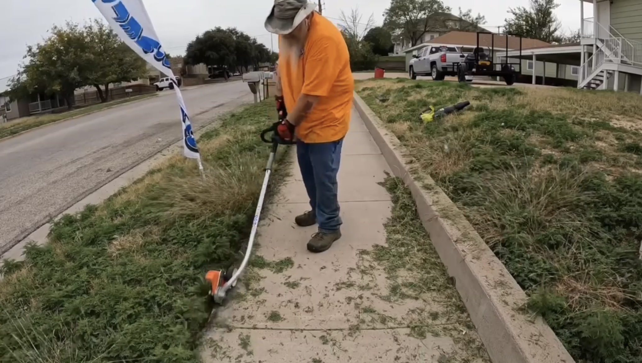 Person in orange shirt and hat using a weed trimmer on a sidewalk, with grass and a street in background.