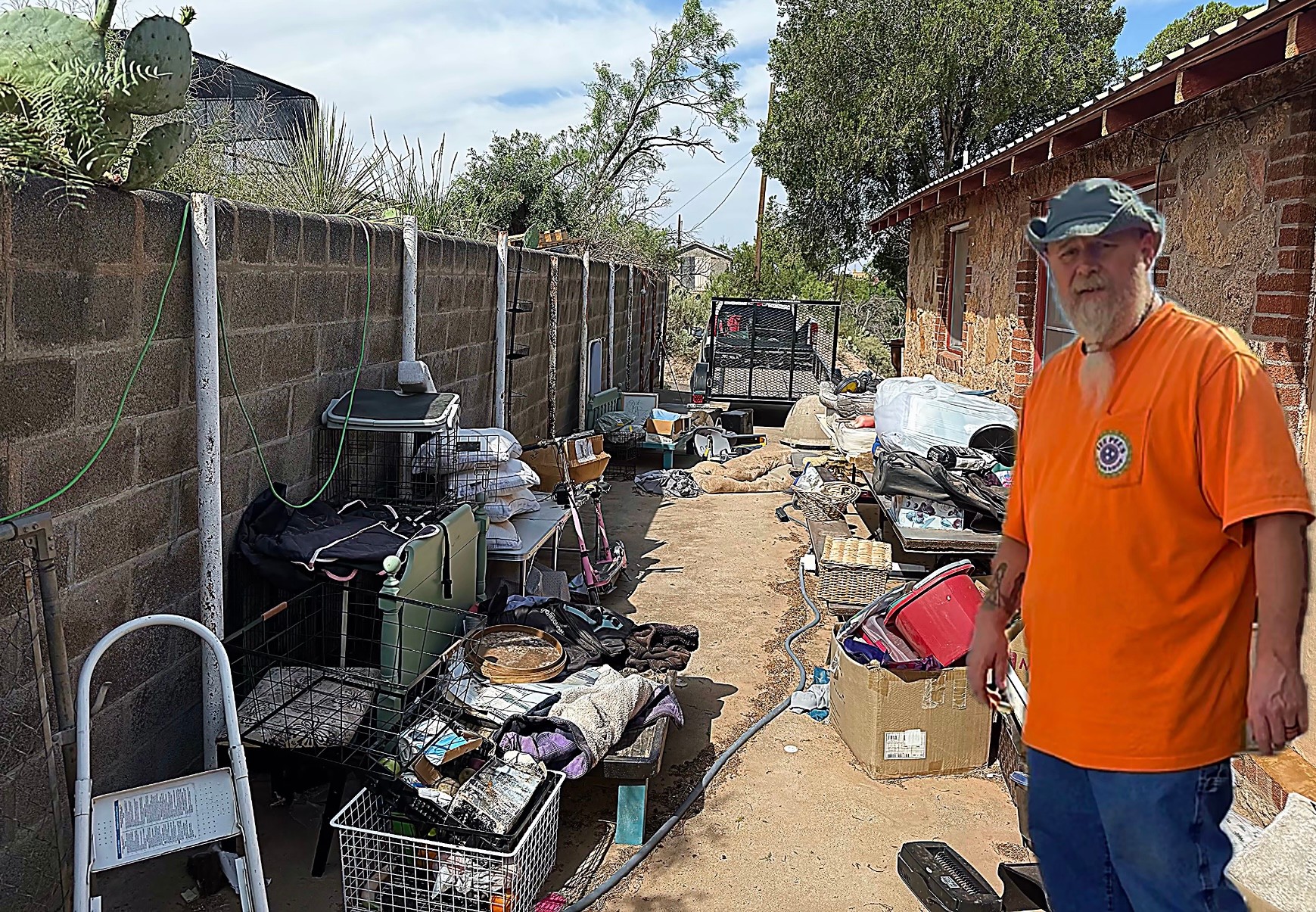 Man in orange shirt and cap standing outdoors near cluttered area with boxes and tools, brick building in background.