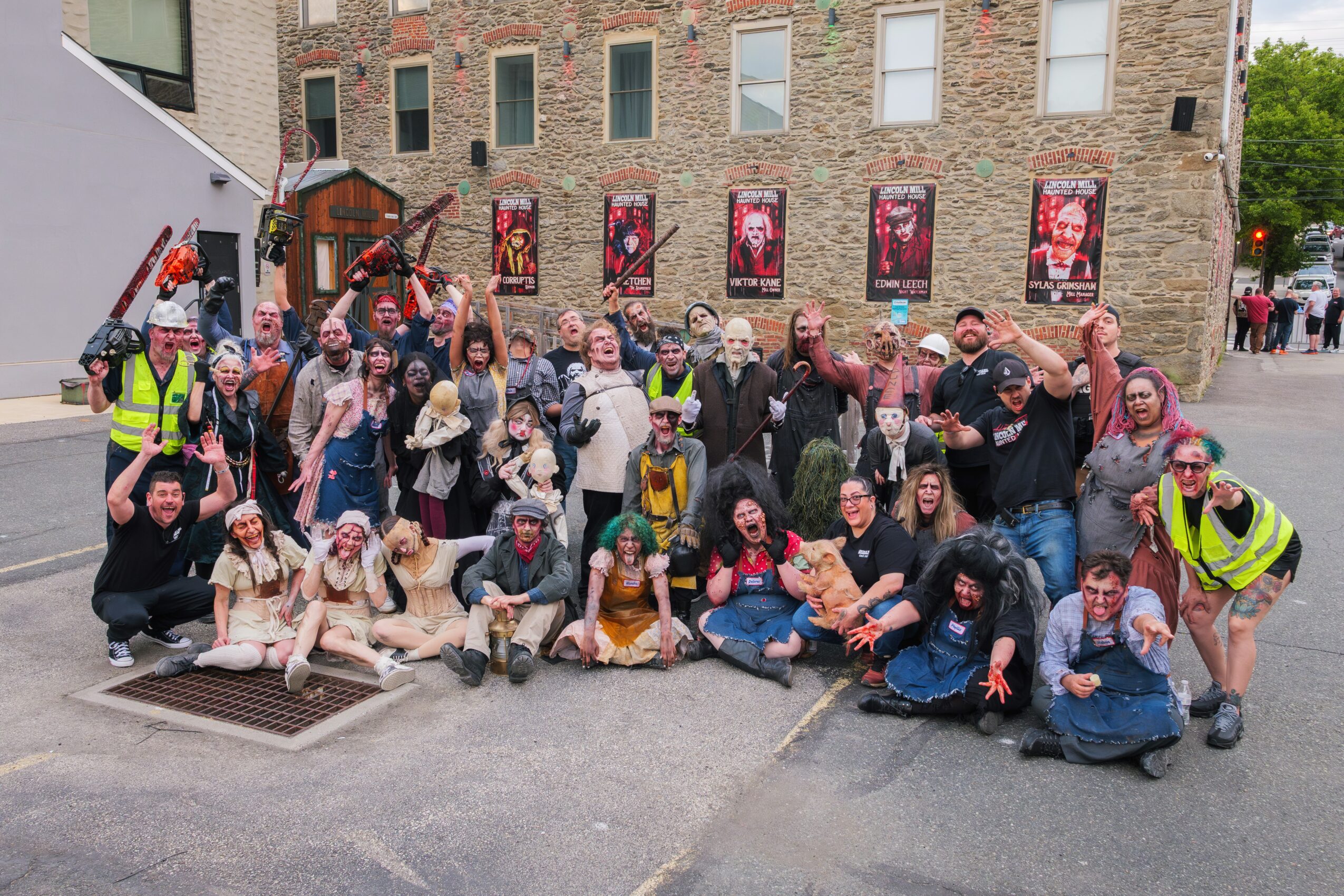 Group of people posing outdoors in front of a brick building, some wearing costumes and safety vests, smiling and making gestures.