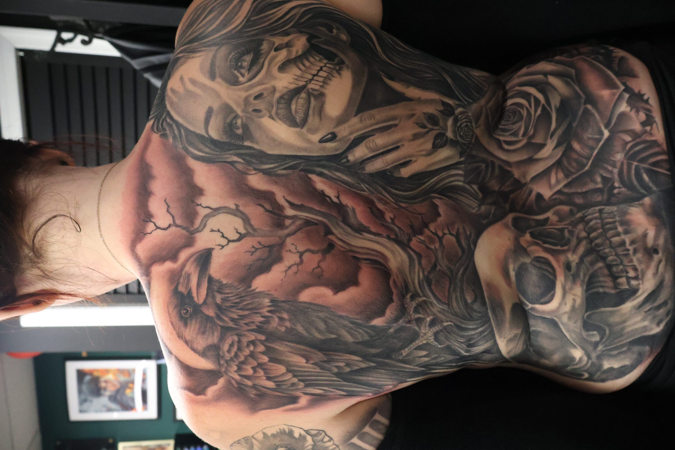 Back view of a person with extensive black and gray tattoos, including a skull, roses, and a dragon, on their back and shoulders.