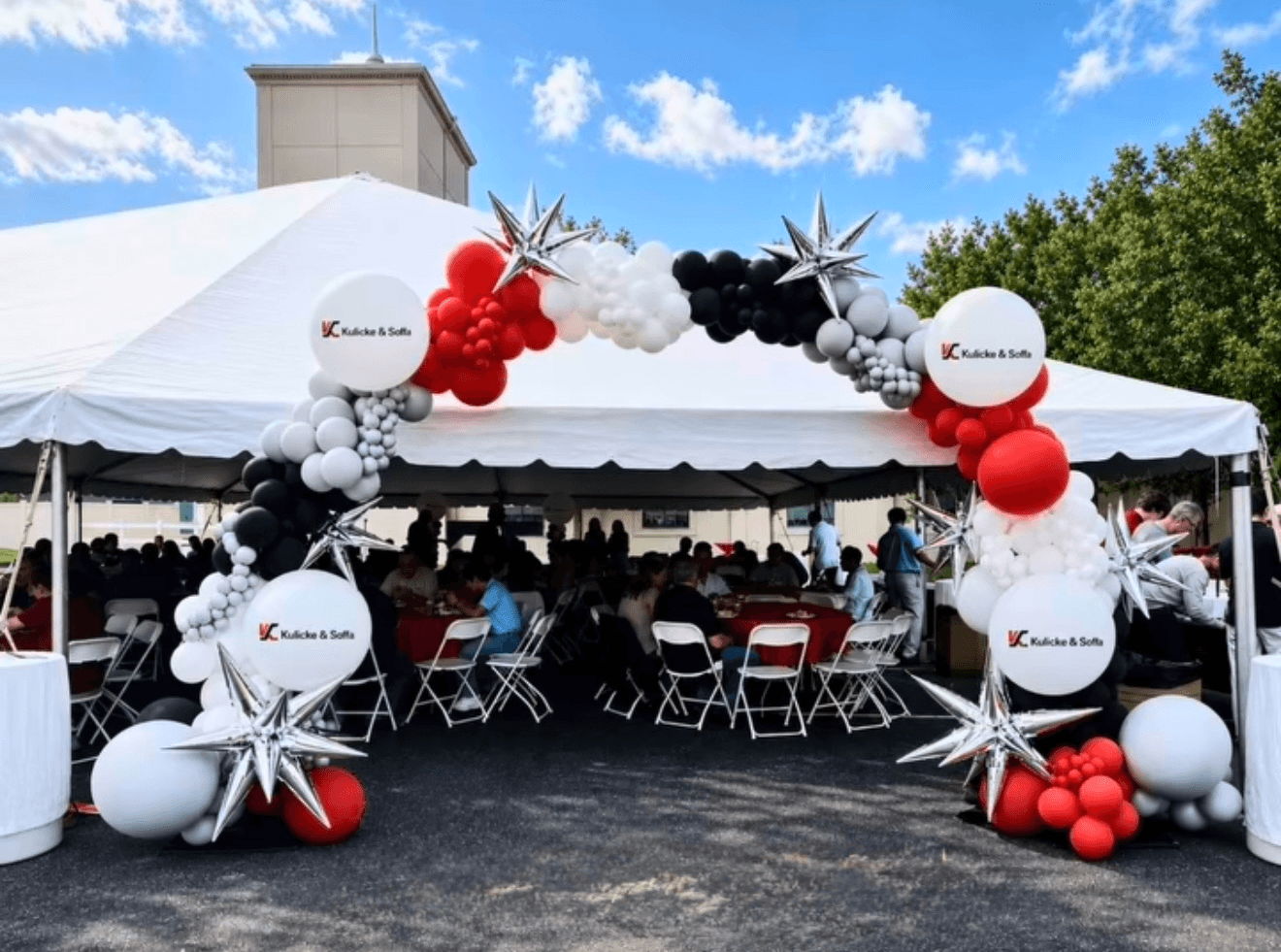 Decorated arch with balloons and stars at outdoor event, tent with seated people underneath, trees and blue sky in background.