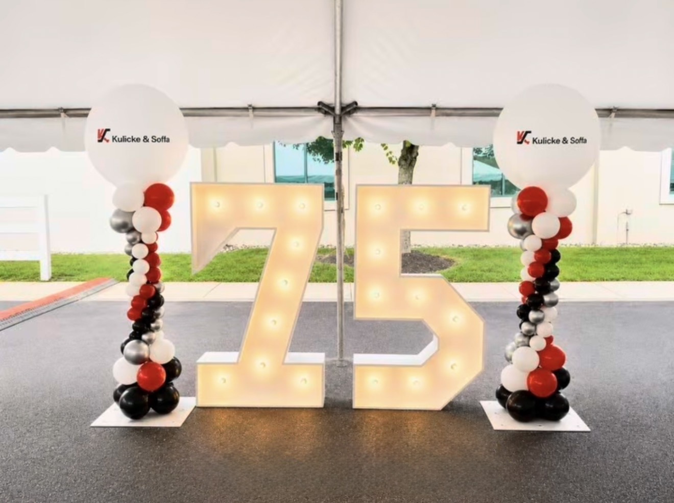 Large illuminated number 75 with balloon columns on each side, inside a tent with white walls and windows.