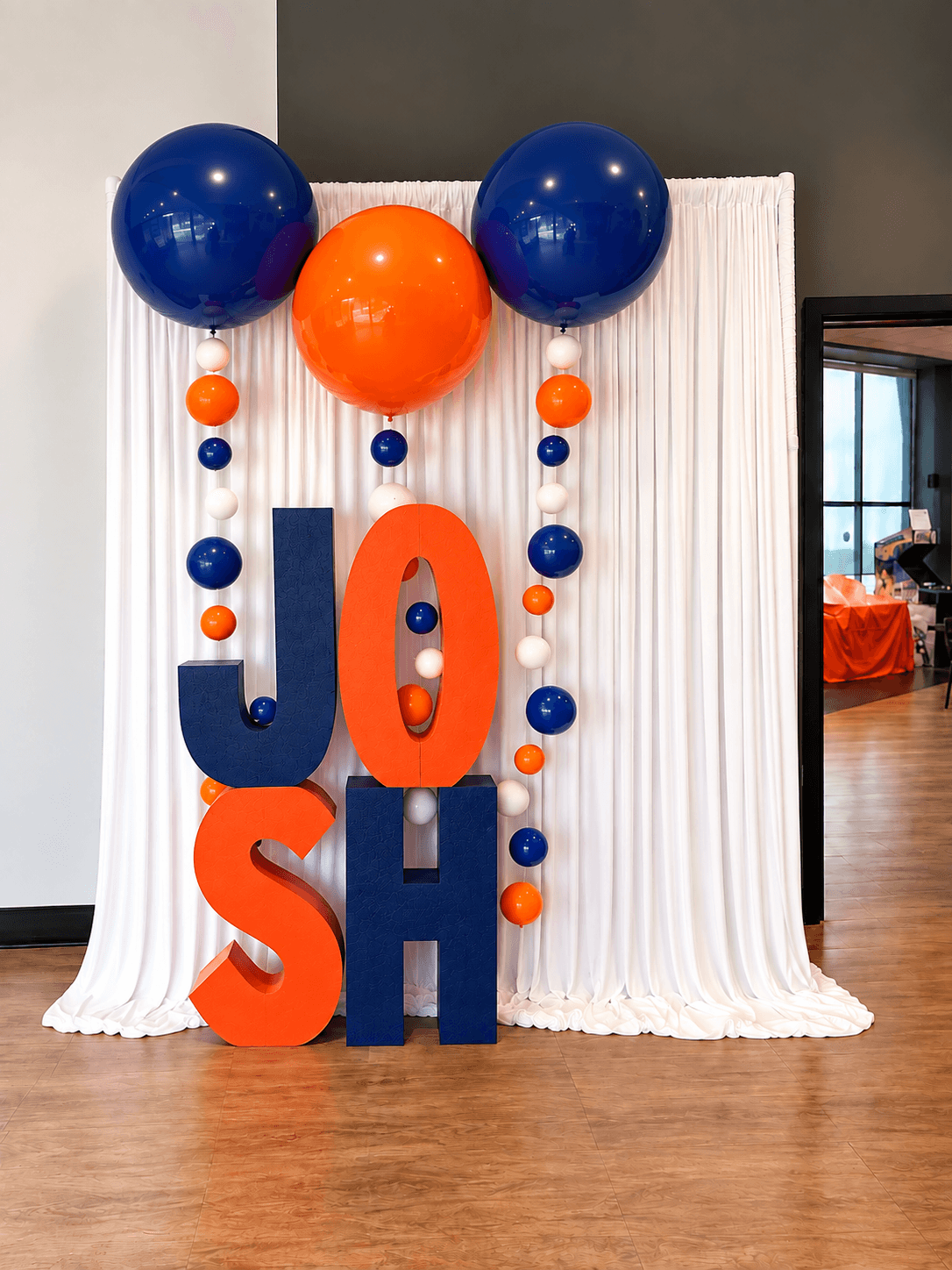 Decorative display with large blue and orange balloons, large orange and blue letters spelling 'JOSH', and a white backdrop.
