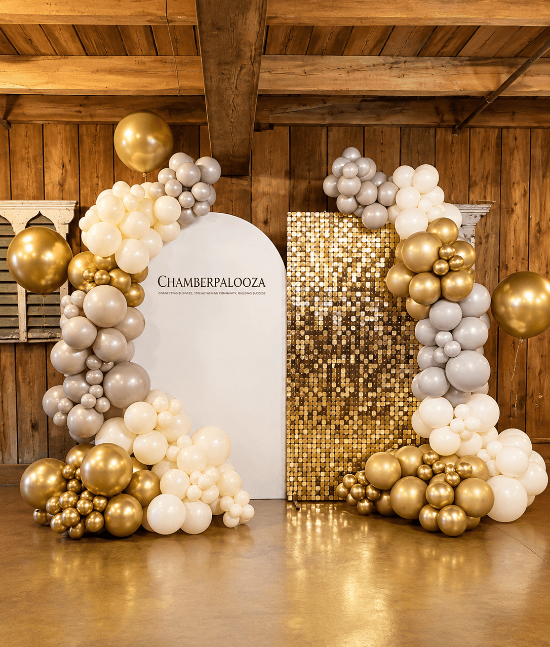 Decorative balloon arch with white, gold, and silver balloons surrounds a white panel and a gold mosaic backdrop, in a wooden room.