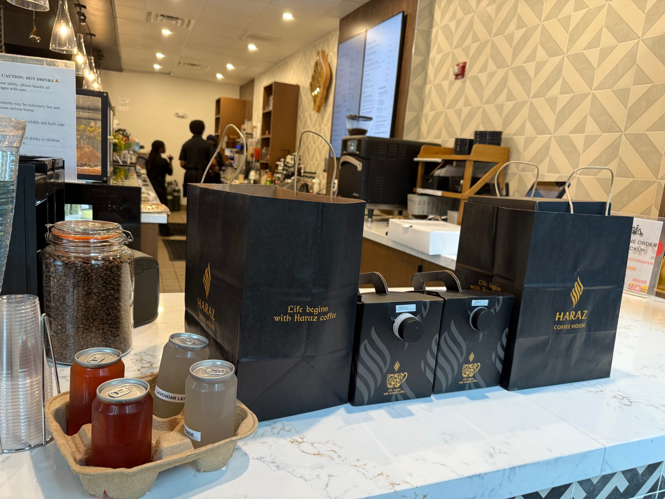 Counter with coffee bags, jars, and cups in a cafe, with people and a patterned wall in the background.
