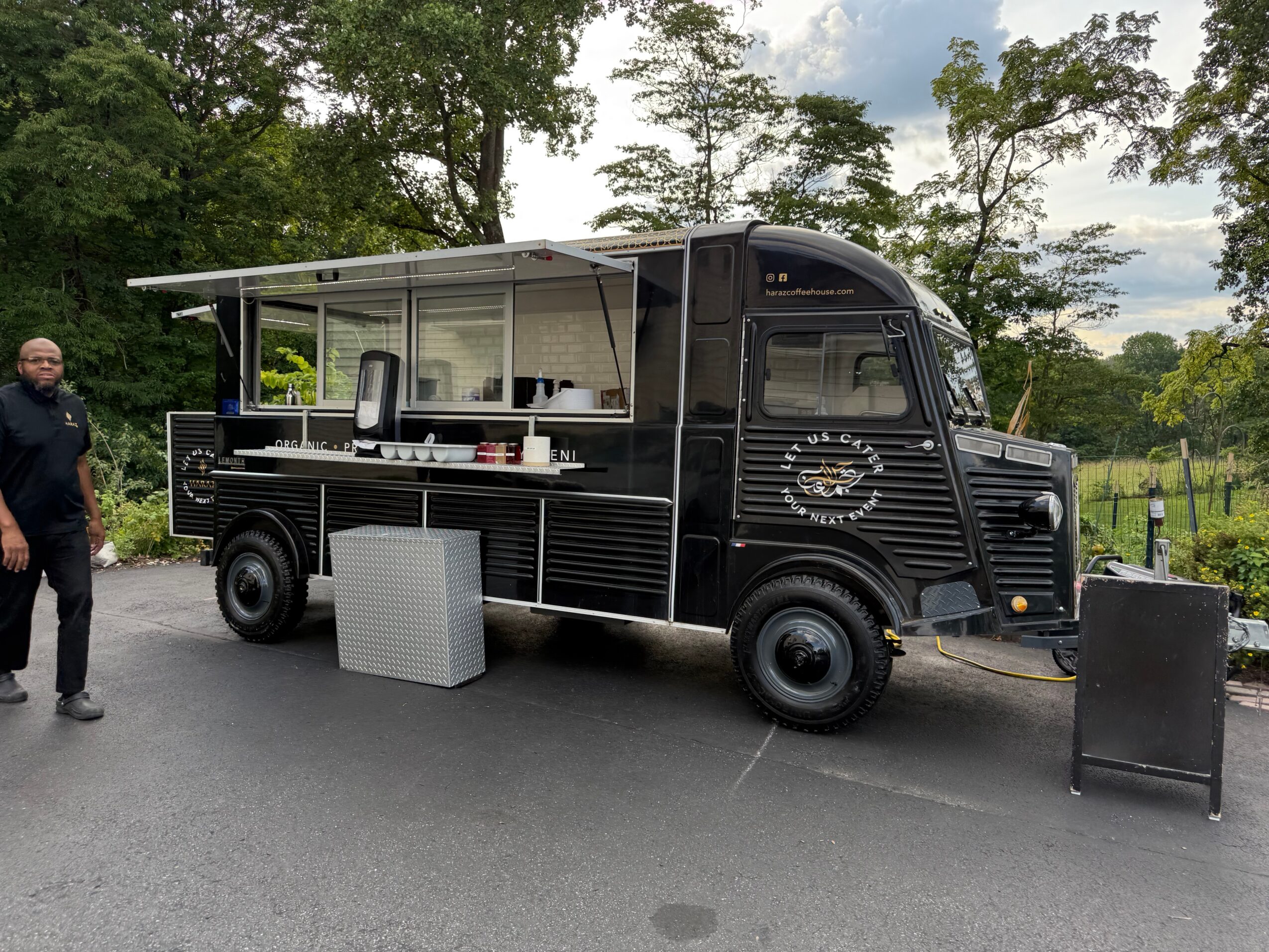 Black food truck with open serving window, outdoor setting with trees, person standing nearby, and various equipment around.