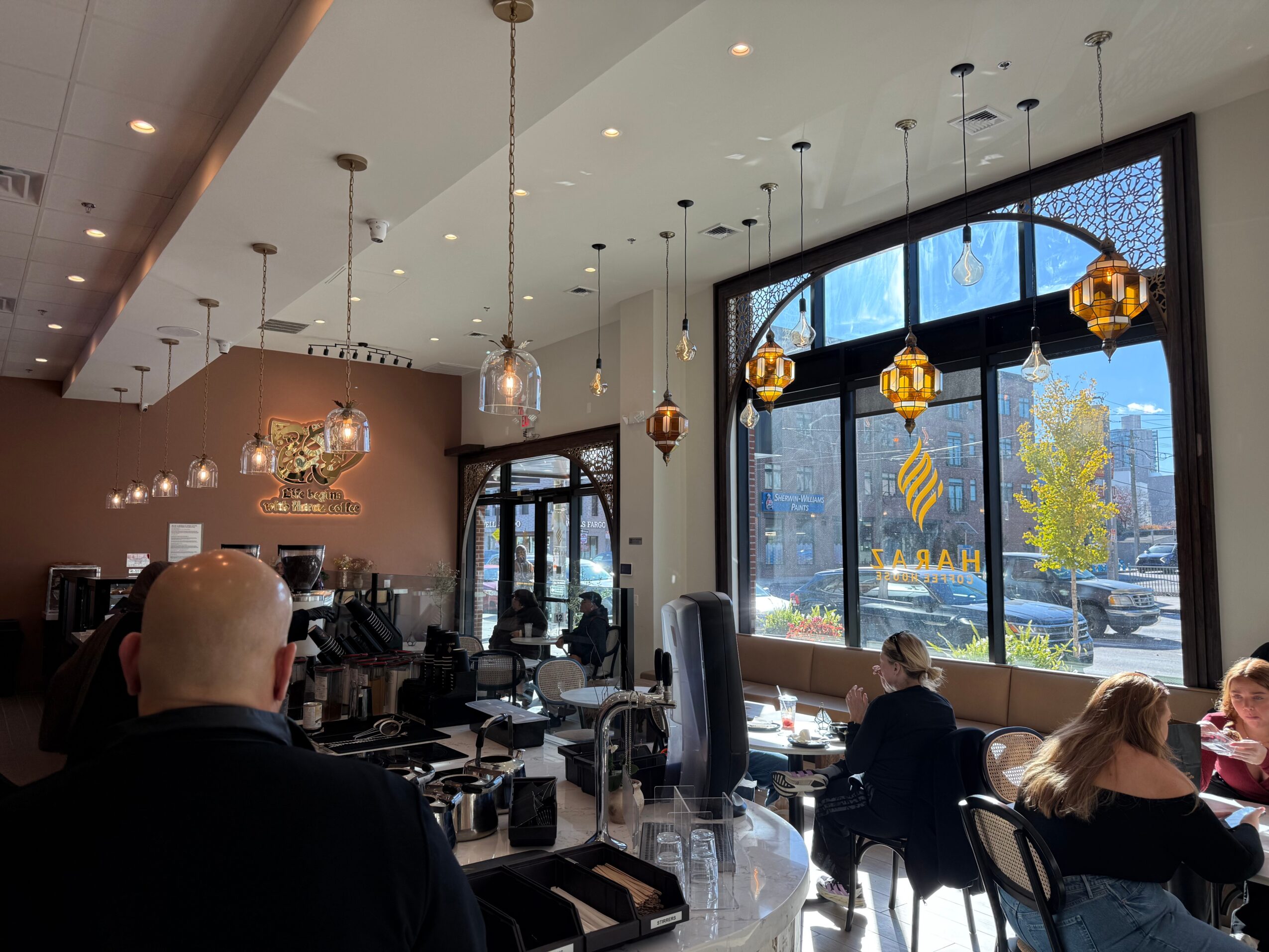Interior of a cafe with hanging pendant lights, large windows, and people sitting at tables.