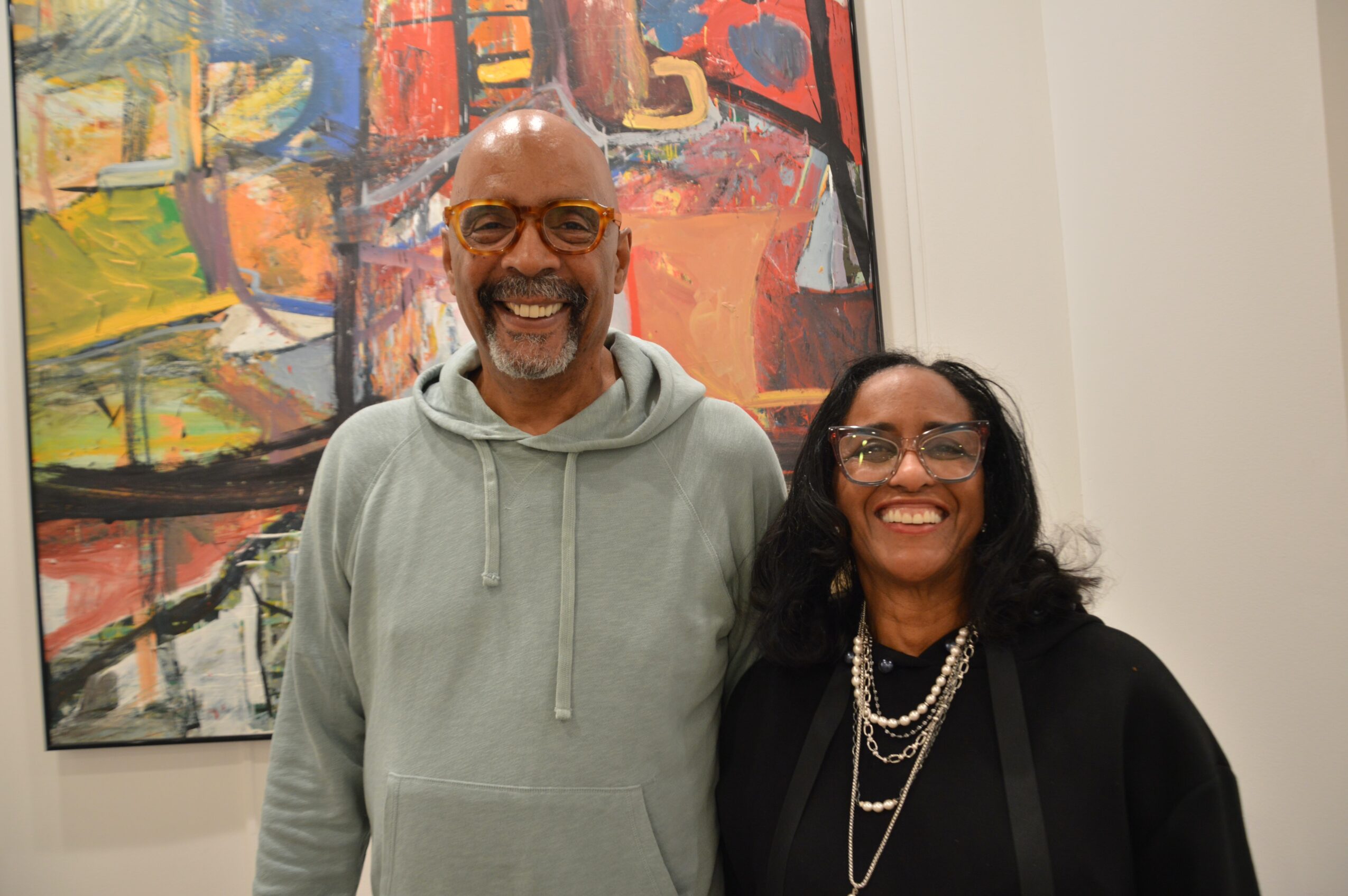 Two smiling people, a man and a woman, standing in front of an abstract painting in an art gallery.