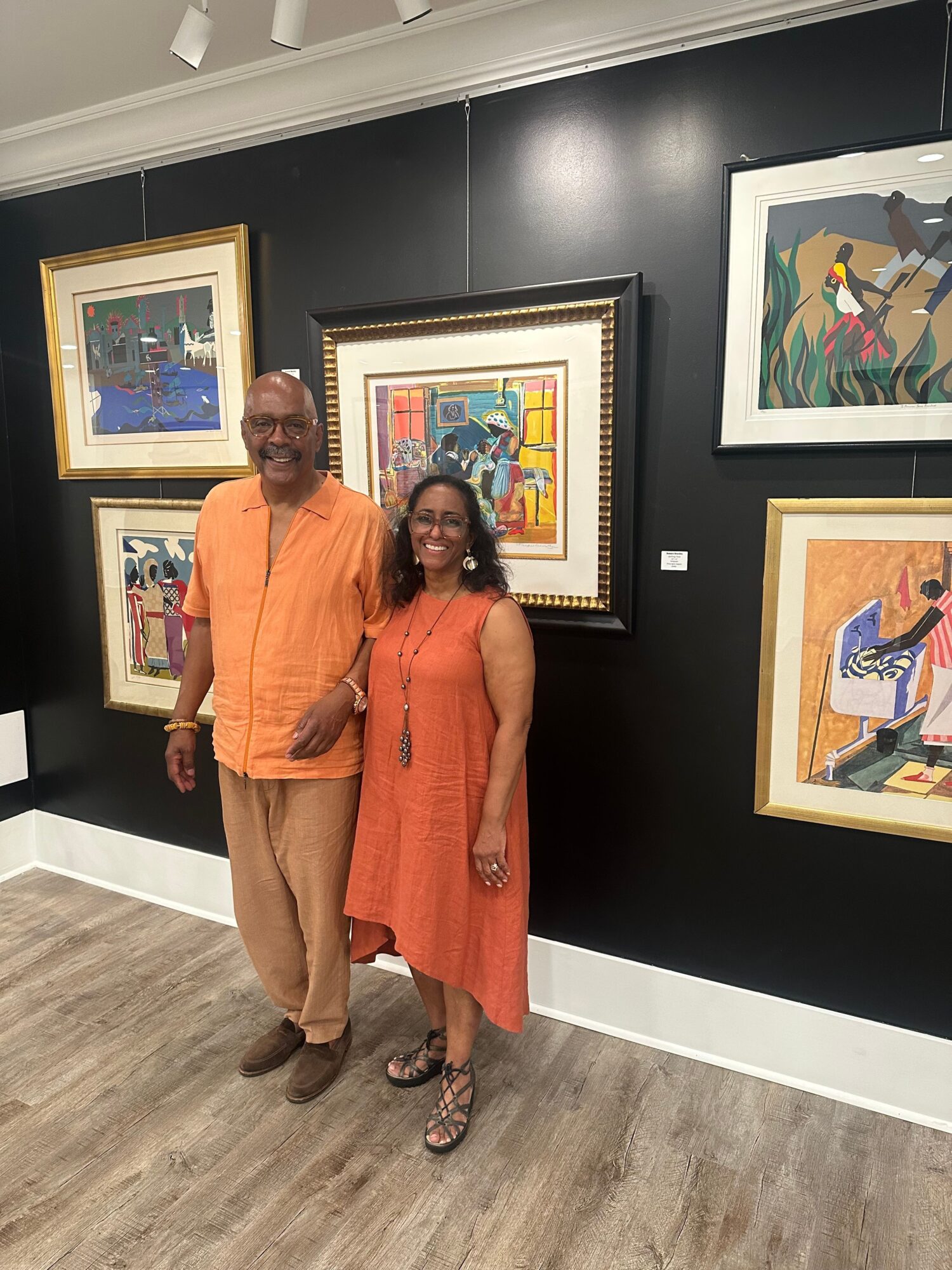 Two people standing in front of framed artwork on a black wall, one wearing an orange dress and the other in orange shirt and brown pants.