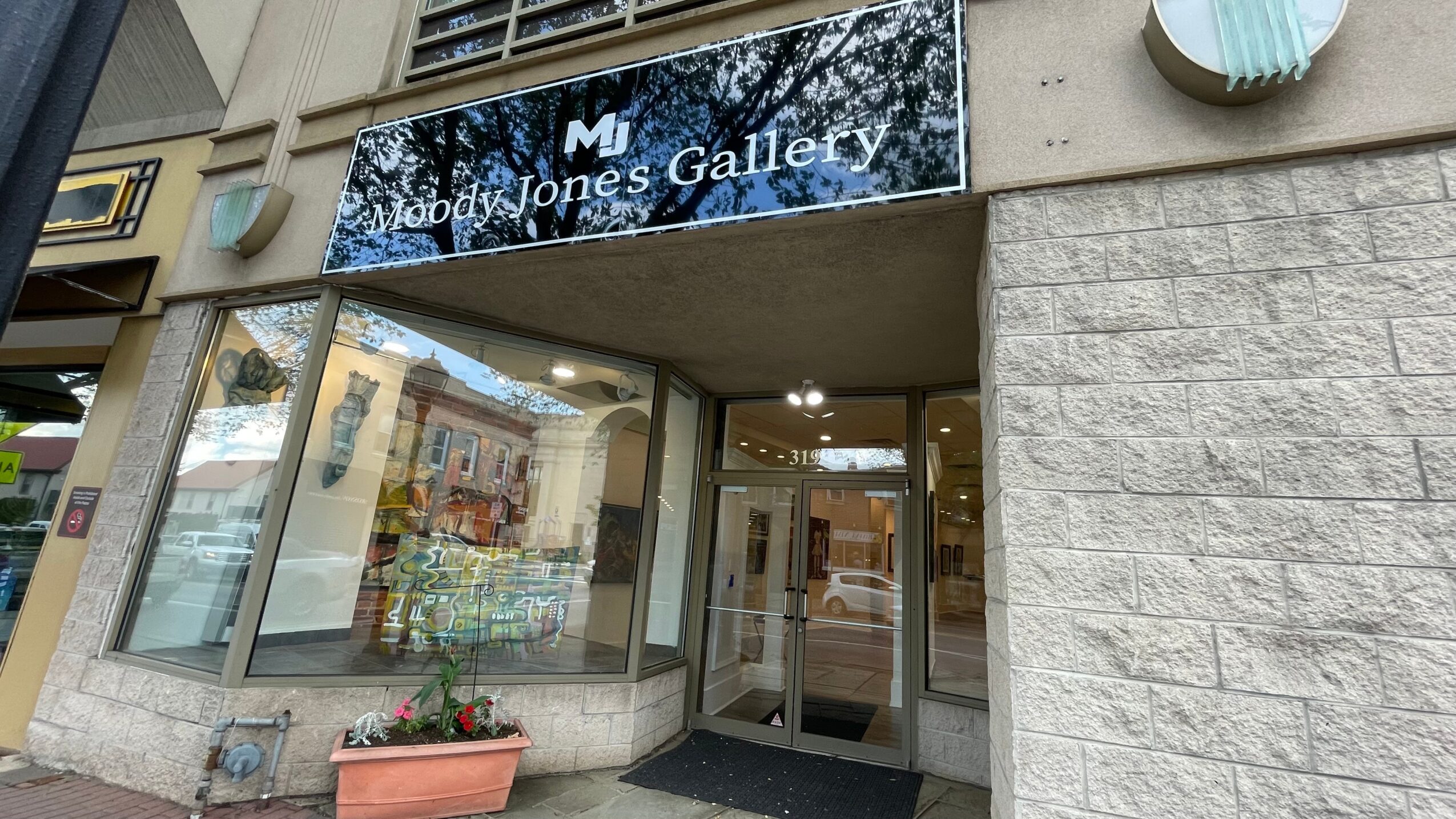 Gallery entrance with glass doors and windows, sign reading 'Moody Jones Gallery,' and a flower pot outside.