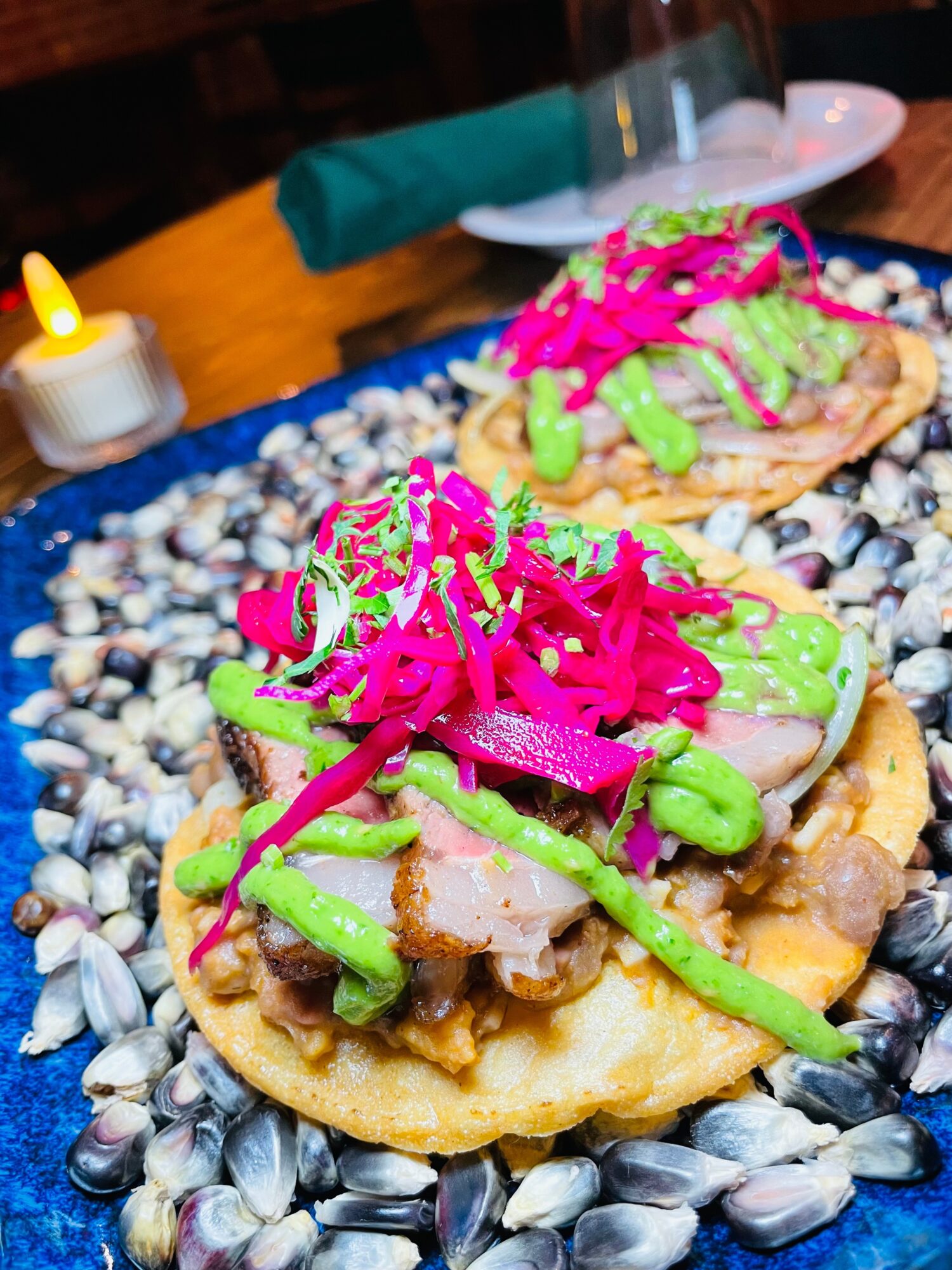Two tacos with meat, green sauce, pink pickled vegetables, on a bed of shells, with a candle and table setting in background.