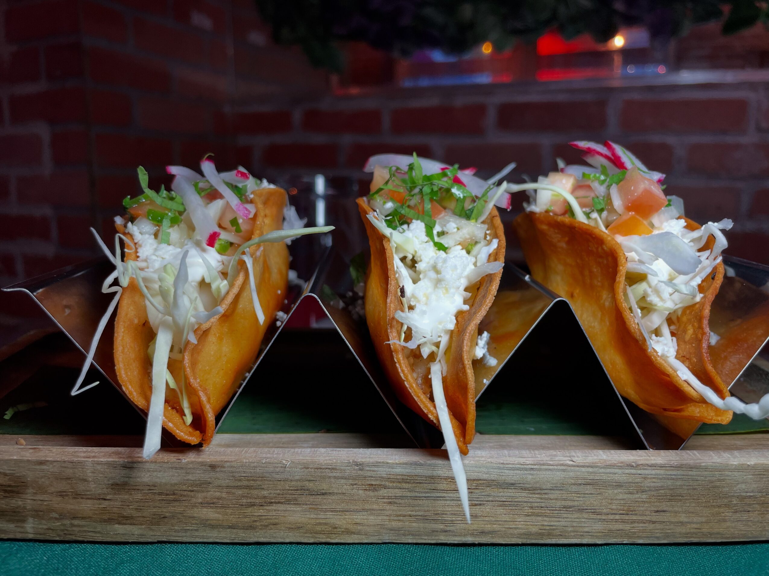 Three tacos filled with shredded meat, lettuce, and cheese on a wooden surface against a brick wall.
