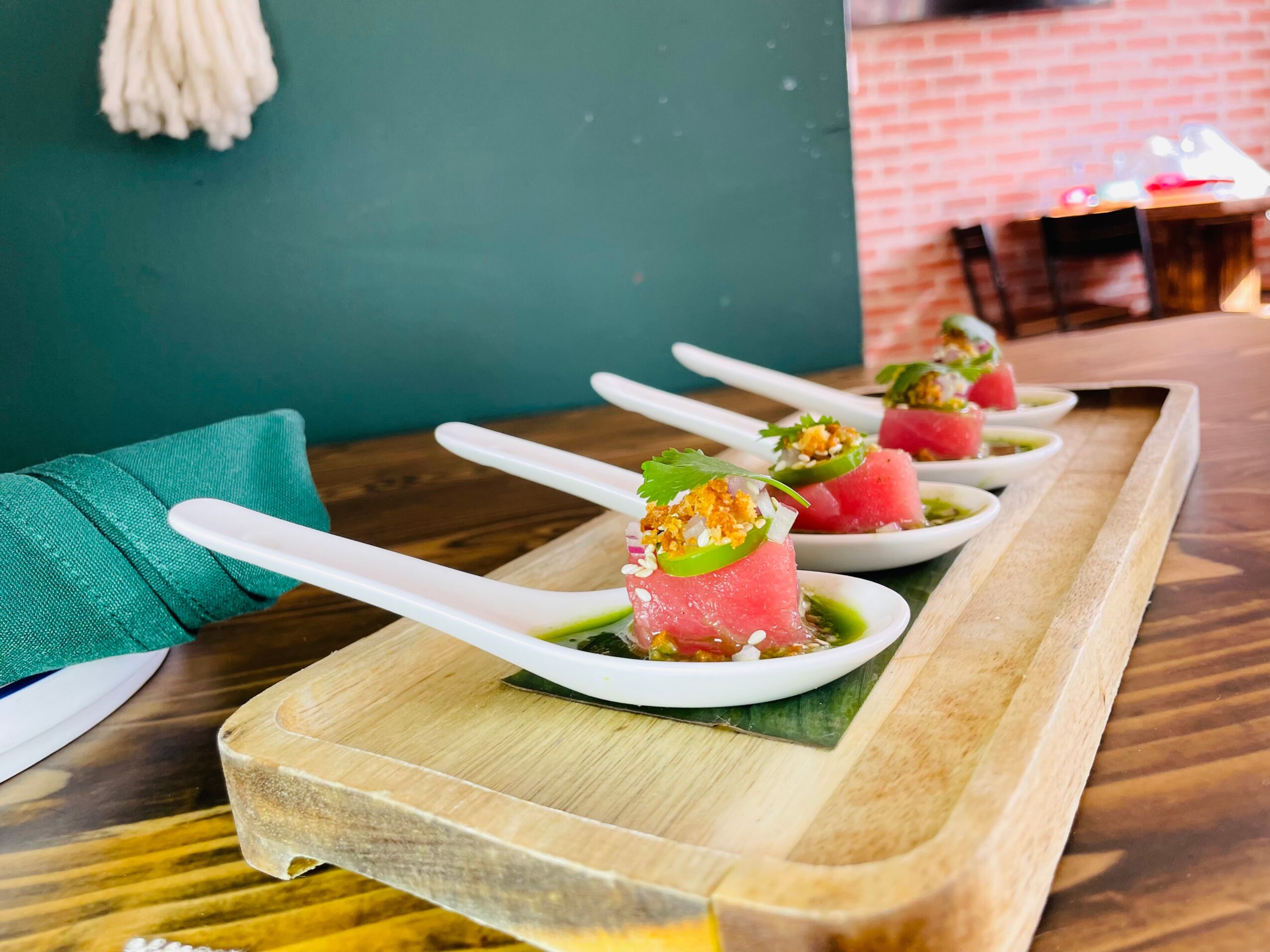 Four small dishes with pink food, garnished with green herbs, on a wooden tray in a restaurant setting.