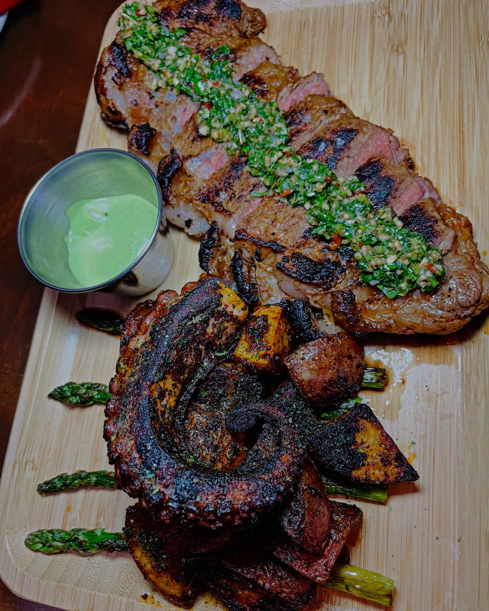 Grilled steak and grilled chicken with green sauce on a wooden board, accompanied by a small green bowl, with sauce drizzled on the meat.