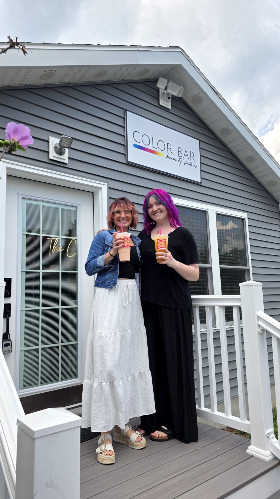 Two women with purple hair holding drinks and smiling outside a building with a sign that reads 'Color Bar'.