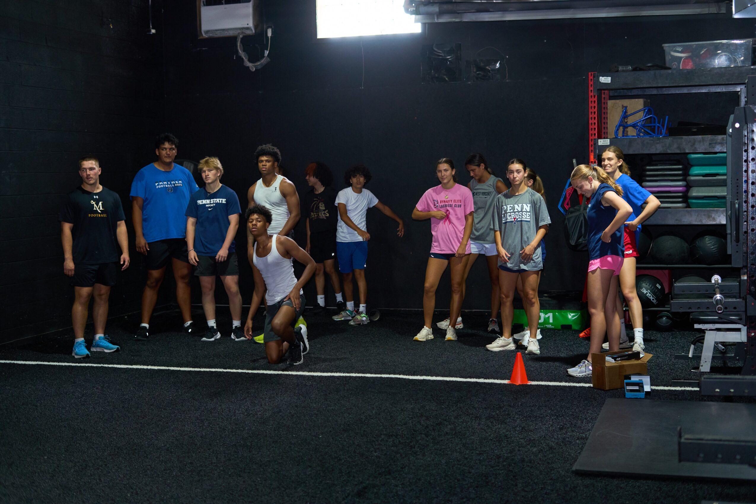 Group of people in a gym, some standing and some preparing for exercise, with equipment and a black wall in the background.