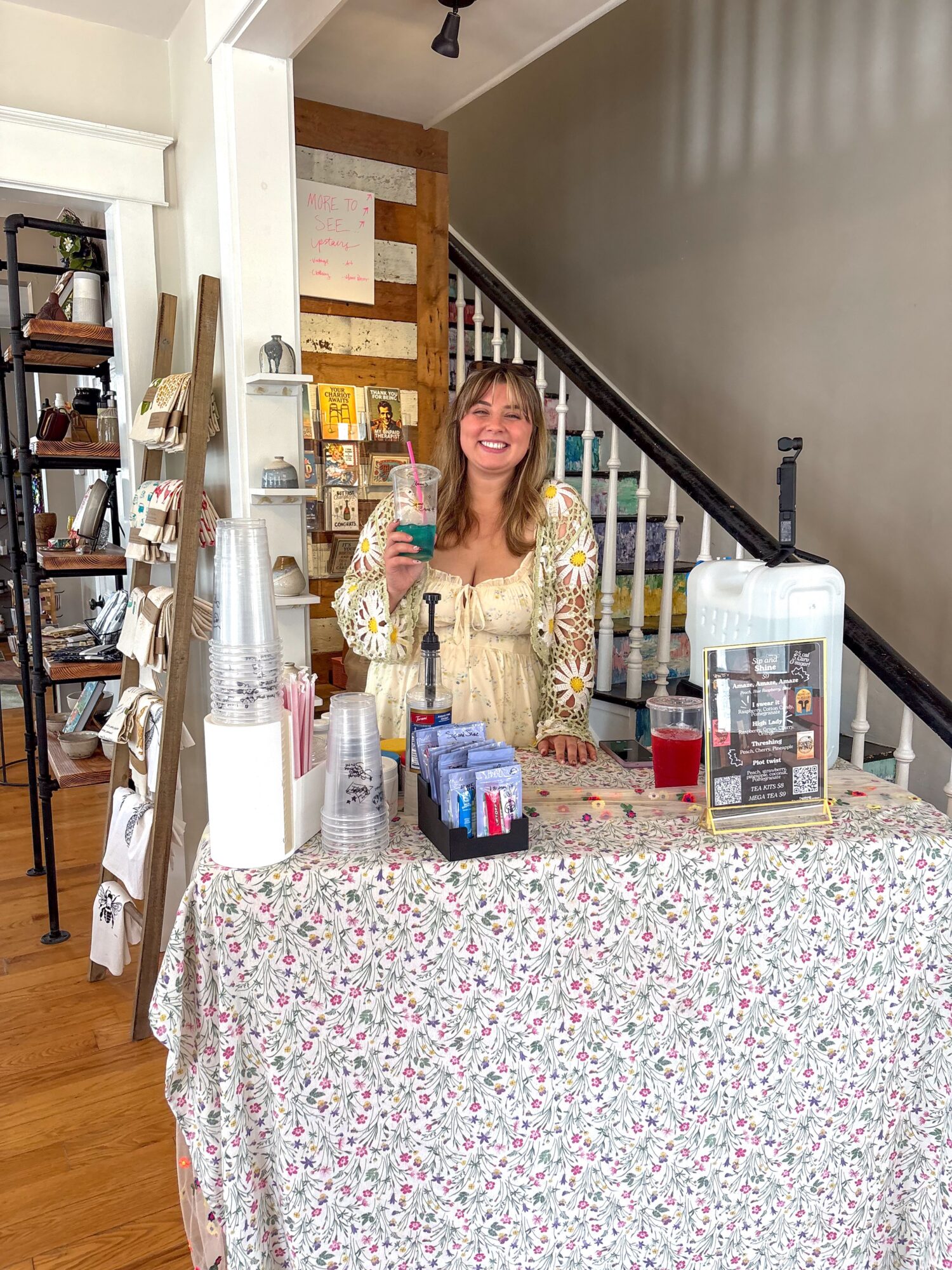 Woman standing behind a table with drinks and snacks, smiling, in a cozy indoor setting.