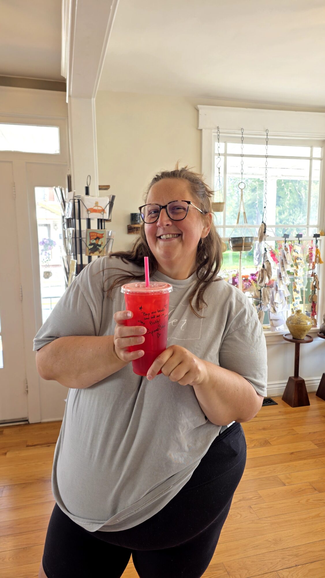 Smiling woman holding a red drink with a straw indoors, with shelves and windows in the background.