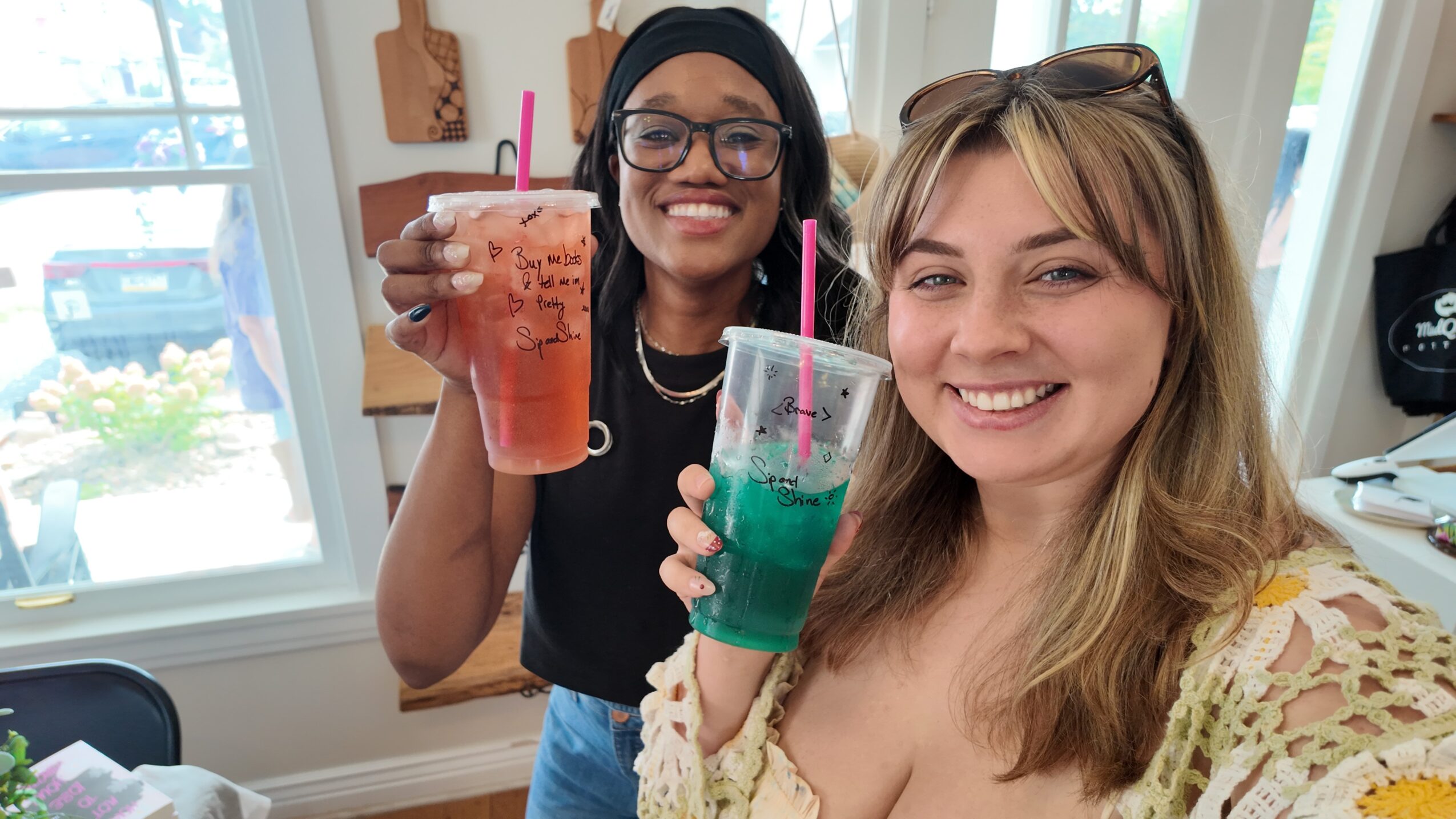 Two women smiling and holding colorful drinks indoors, with windows and a bright room in the background.