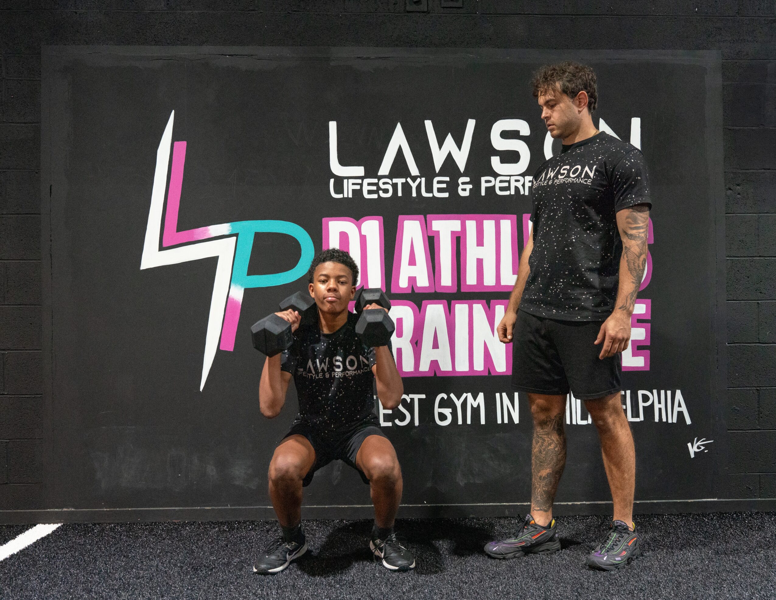 Two men in athletic clothing pose in front of a black wall with colorful text and logo, one squatting and one standing.