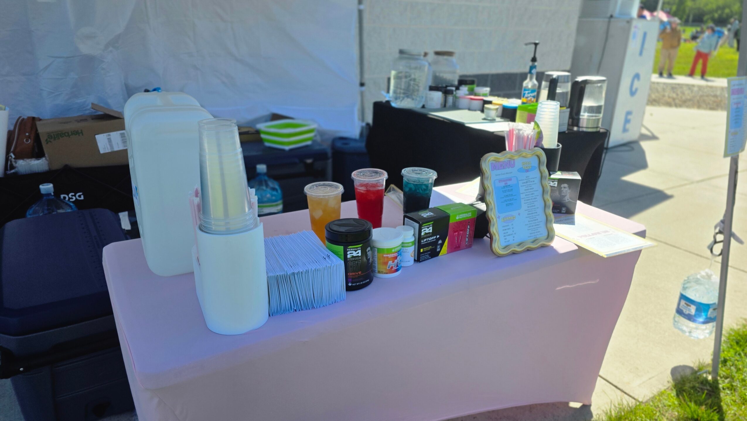 Table with cups, containers, and a framed sign at an outdoor event, sunlight, and tents in background.