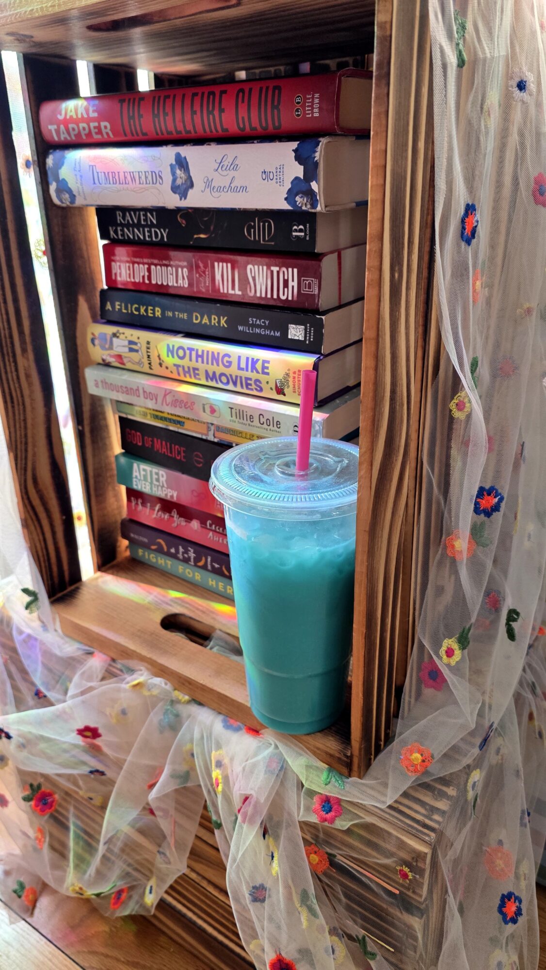 A blue drink with a pink straw on a wooden shelf, with books behind and colorful fabric in front.