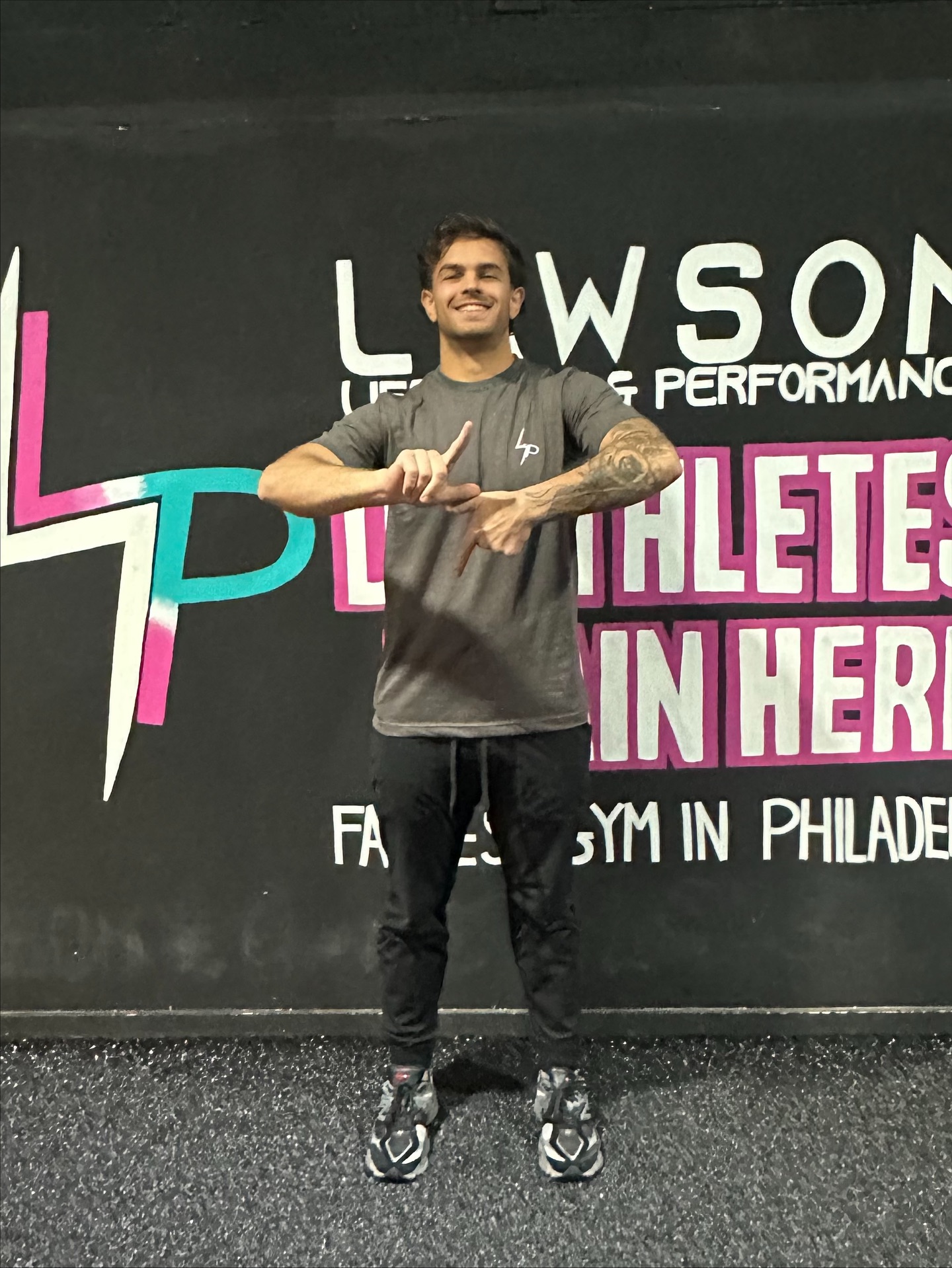 Young man with tattoos smiling, standing in front of a black wall with colorful text, wearing a gray shirt and black pants.