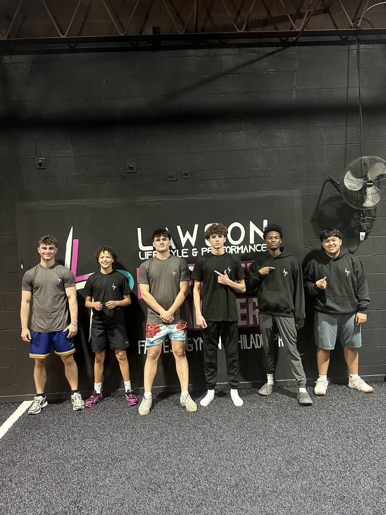Group of seven young people standing in a line indoors, some with hands in pockets, in front of a black wall with text.