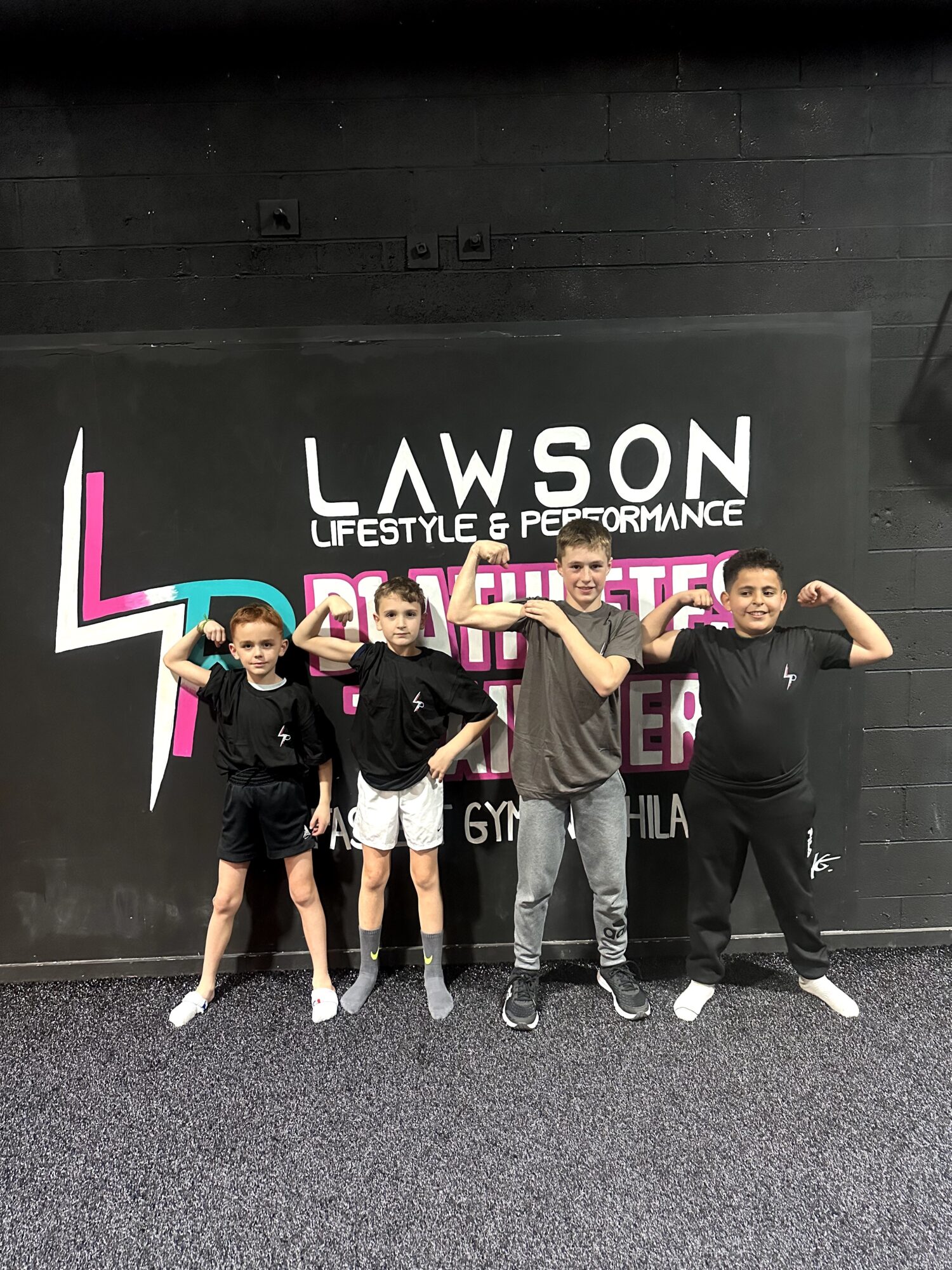Five children standing in front of a black wall with a Lawson sign, some flexing muscles, in a gym setting.
