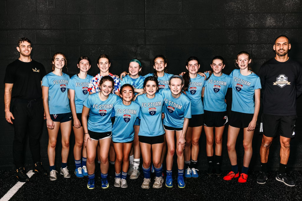 Group of young female athletes in blue sports uniforms with two adult coaches, standing on a dark indoor court, smiling.
