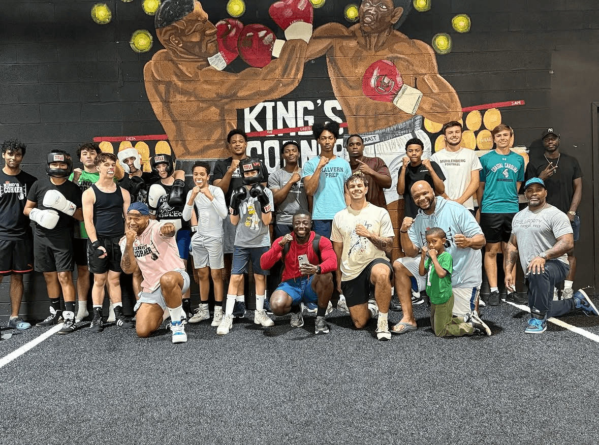Group of people posing in front of a boxing-themed mural with fighters and boxing gloves, some wearing masks, in a gym or sports venue.