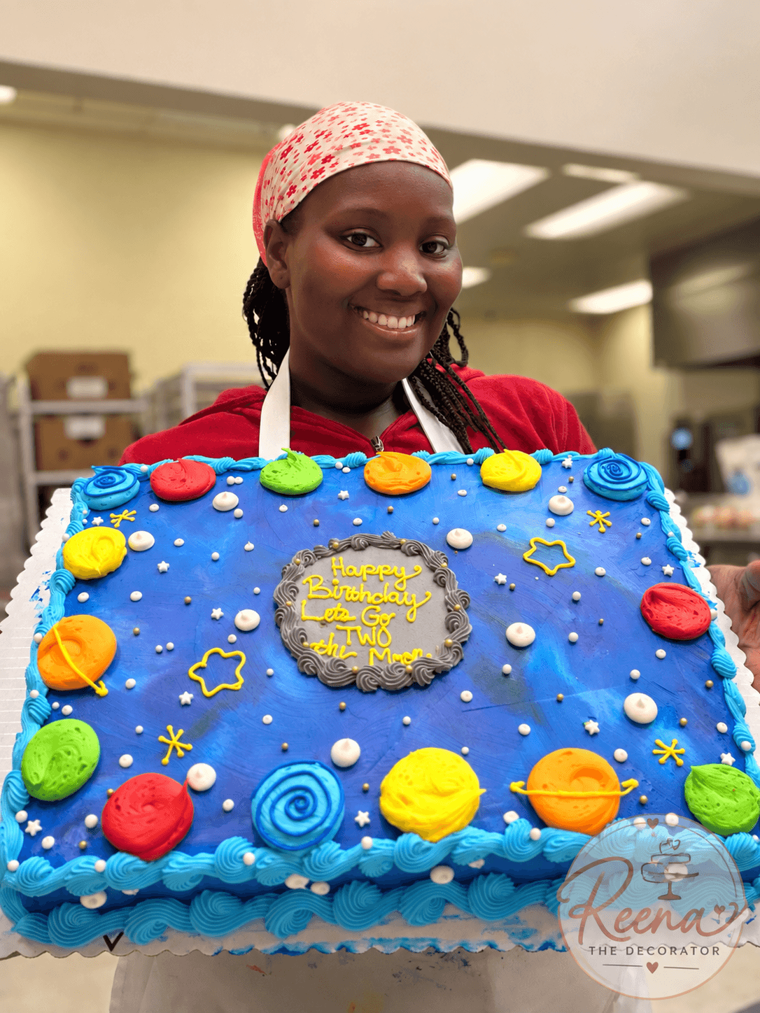 Smiling woman holding a decorated birthday cake with planets and stars, in a room with shelves and lighting, wearing a pink headscarf.