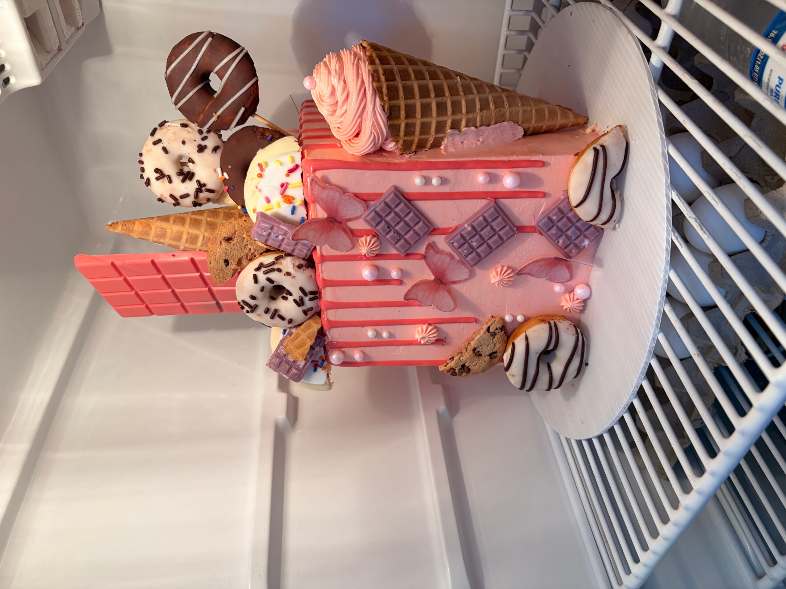 Ice cream cone, chocolate, and cookie desserts on a pink cake with candies and cookies, on a white surface.